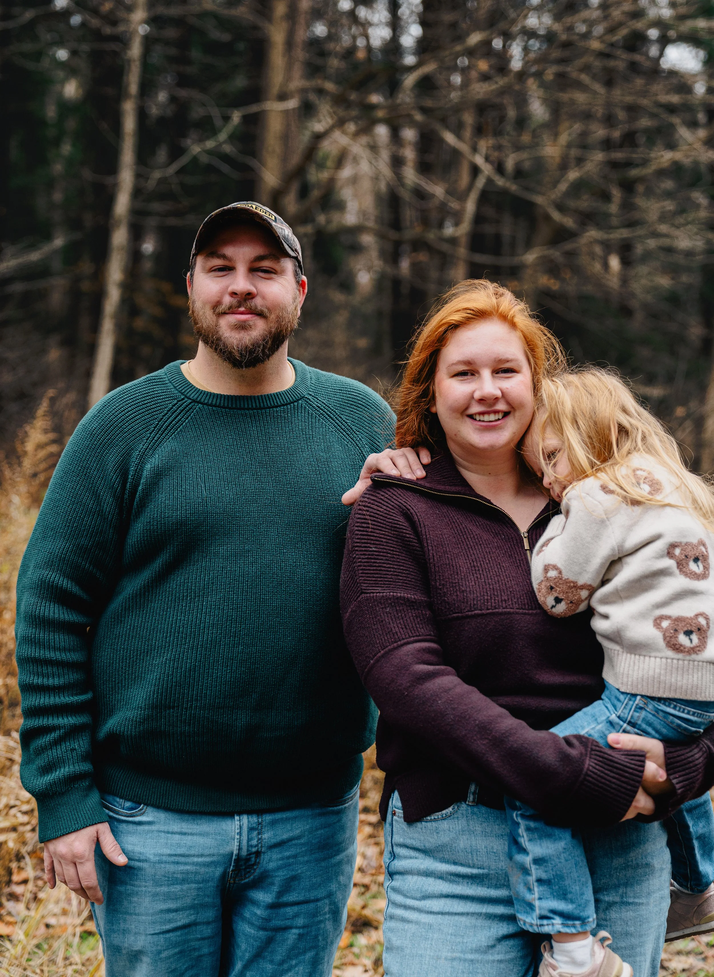 A family of three outdoors in a wooded area during autumn, with a man wearing a green sweater and a camouflage cap, a woman holding a young girl with red hair and a bear-patterned sweater.