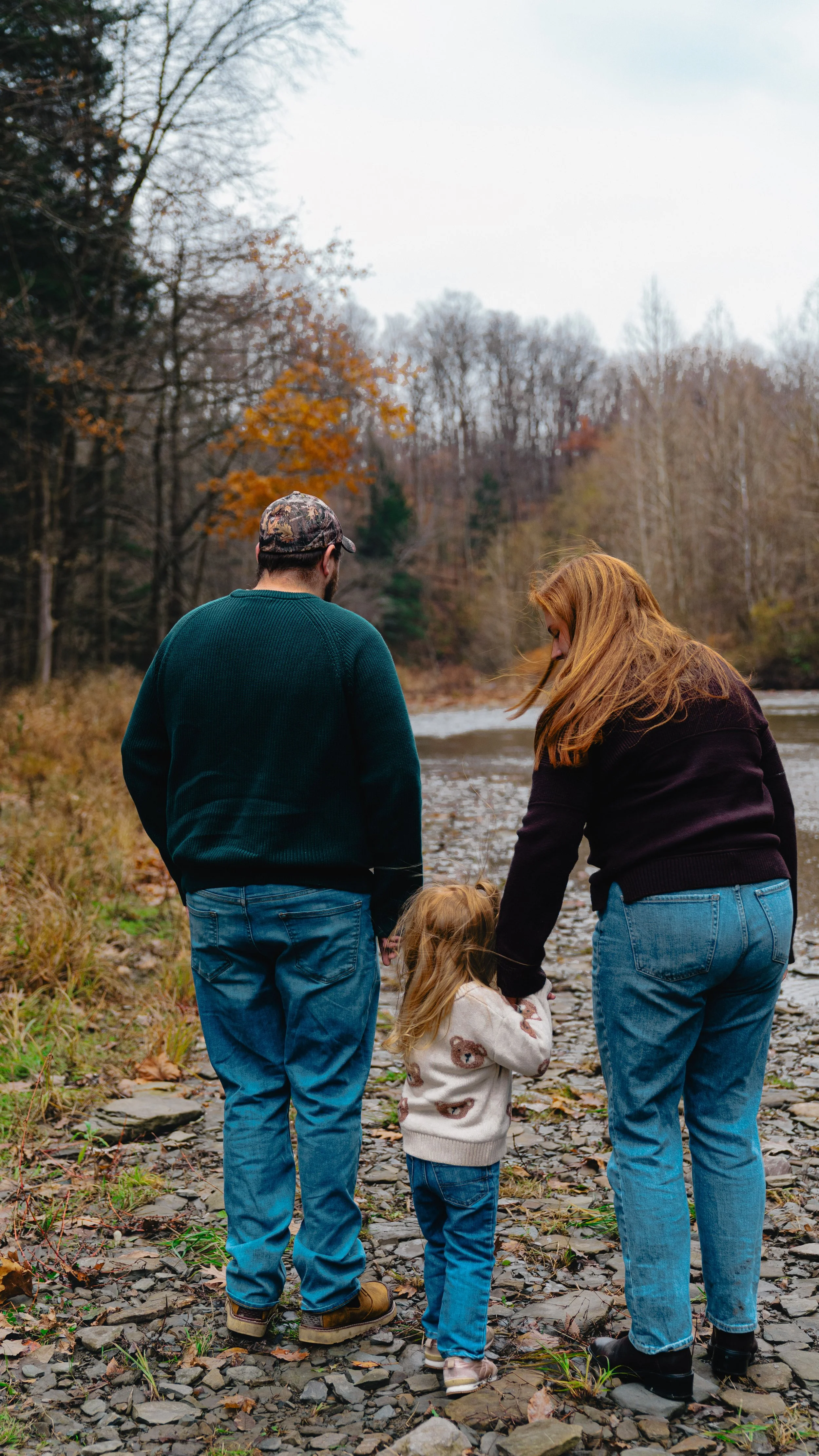 A family of three, two adults and a young girl, holding hands and walking near a river on a rocky shoreline during fall with trees in the background.