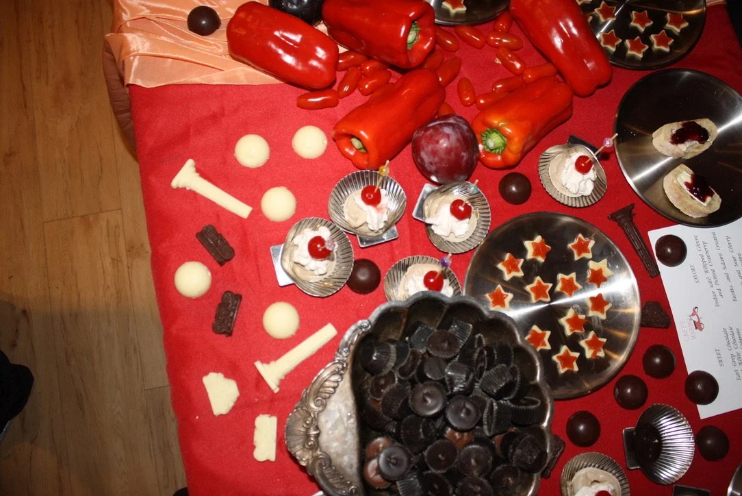 A table with various chocolates, red bell peppers, tomatoes, grapes, and cupcakes decorated with cherries on top. There are also star-shaped cookies on a silver plate and some candy corns. The table is covered with a red tablecloth.