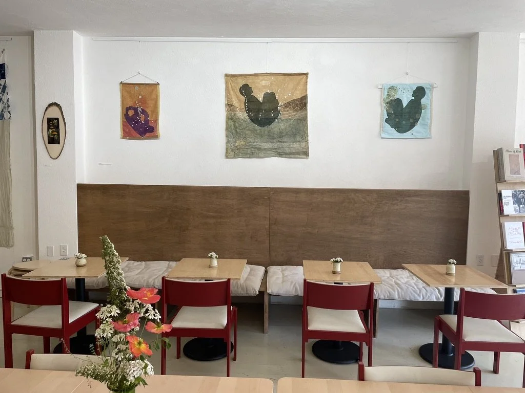 Interior of a cafe or restaurant with wooden and red chairs, small tables, flower vases, and hanging artwork on the white walls.
