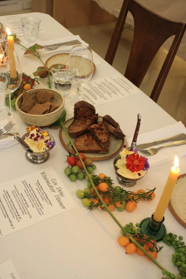A festive dinner table set with bread, grilled vegetables, pudding with edible flowers, and a candle, decorated with tomatoes and a flowering vine.