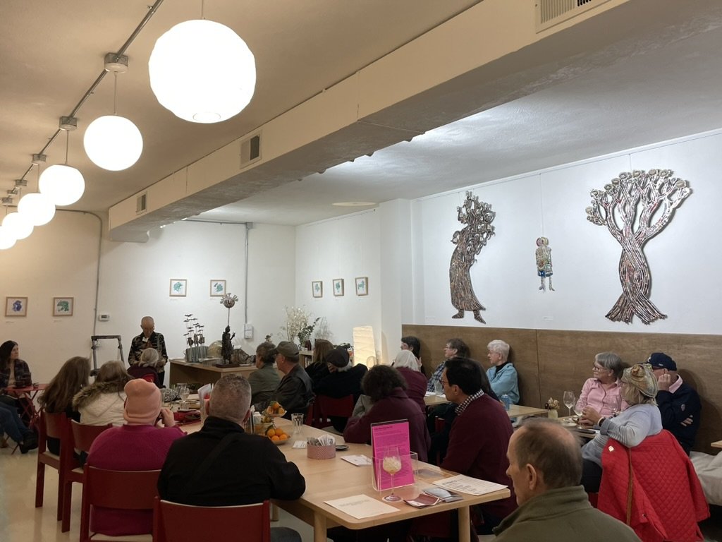 A group of people sitting at tables in a restaurant or cafe, listening to a person speaking at the front. The room has white walls with decorative art and hanging globe lights.