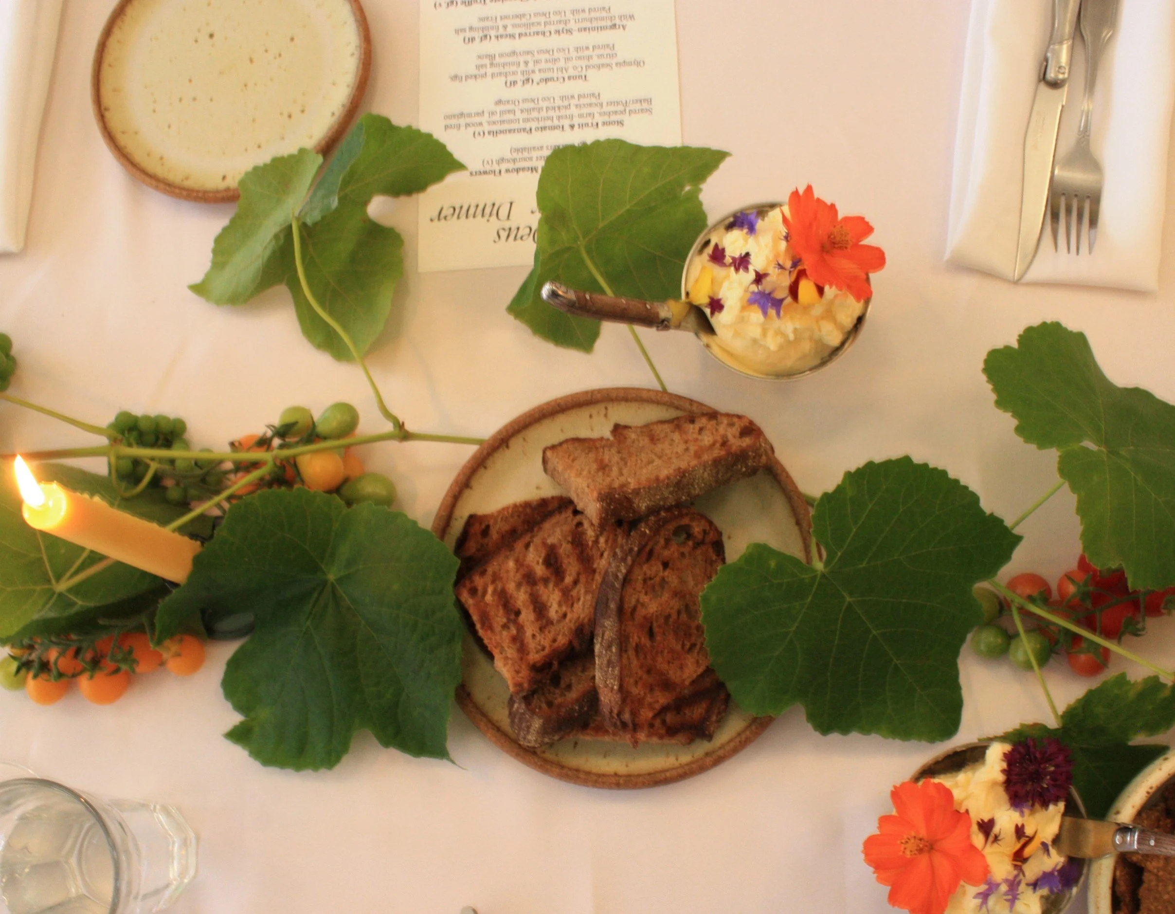 A white table setting featuring a small bowl of salad, slices of grilled meat, and a bowl with edible flowers. The table is decorated with green leaves and small orange berries, and a lit yellow candle is on the side. Silverware is arranged on either