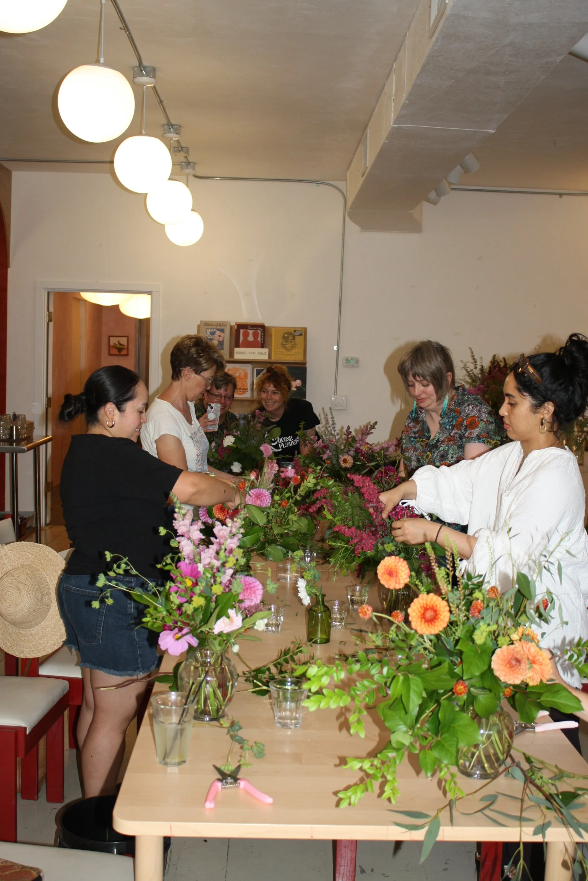 Women arranging colorful flower bouquets on a long table in a room with beige walls and ceiling lights.