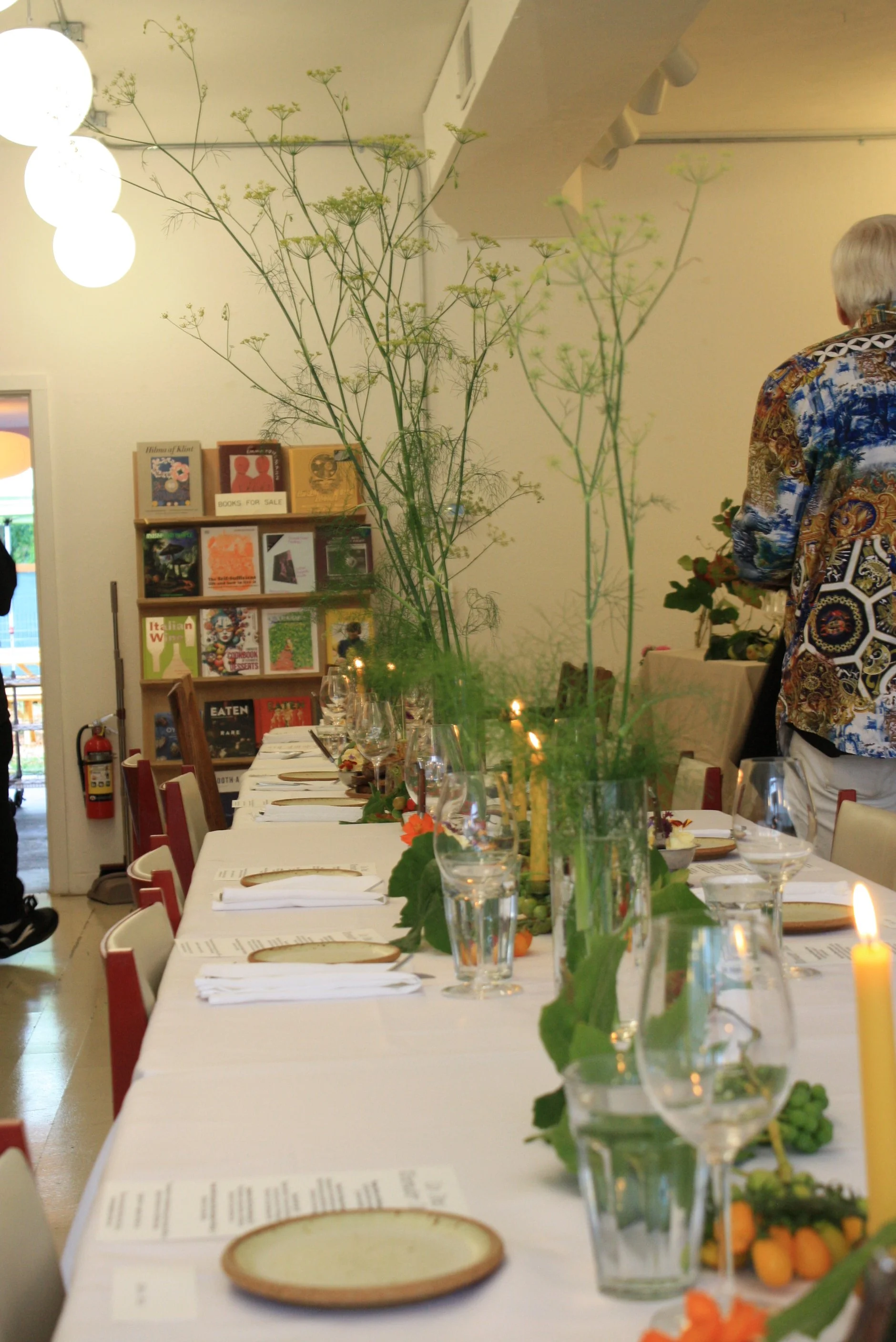 A long dining table set for a meal with plates, glasses, and napkins, decorated with tall green vegetable arrangements. In the background, bookshelves and a person wearing a patterned Hawaiian shirt are visible inside a well-lit room.