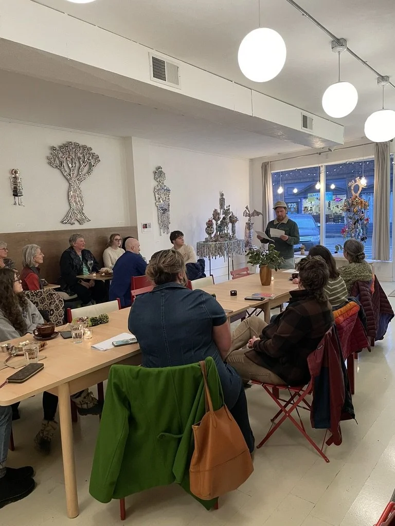 A man giving a speech or reading to a seated audience in a cozy indoor space with artsy wall decorations, including metal trees and a display of decorative objects, during evening time visible through the window.
