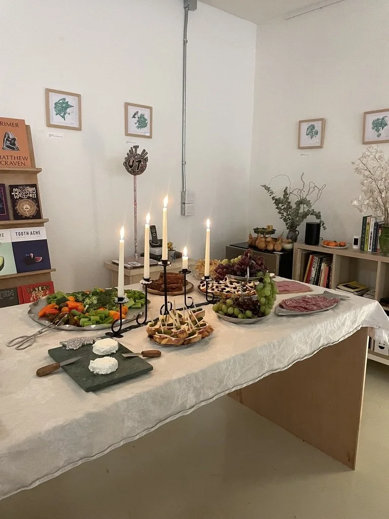 A table set for a celebration with candles, assorted vegetables, grapes, cheese, and a meat platter in a cozy room with books and framed pictures on the wall.