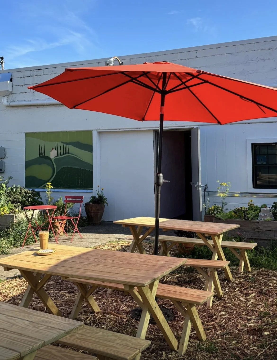 Outdoor patio scene with wooden picnic tables and benches, a red patio umbrella, potted plants, a small pink metal table and chair set, and a building with a painting of a landscape on the wall.