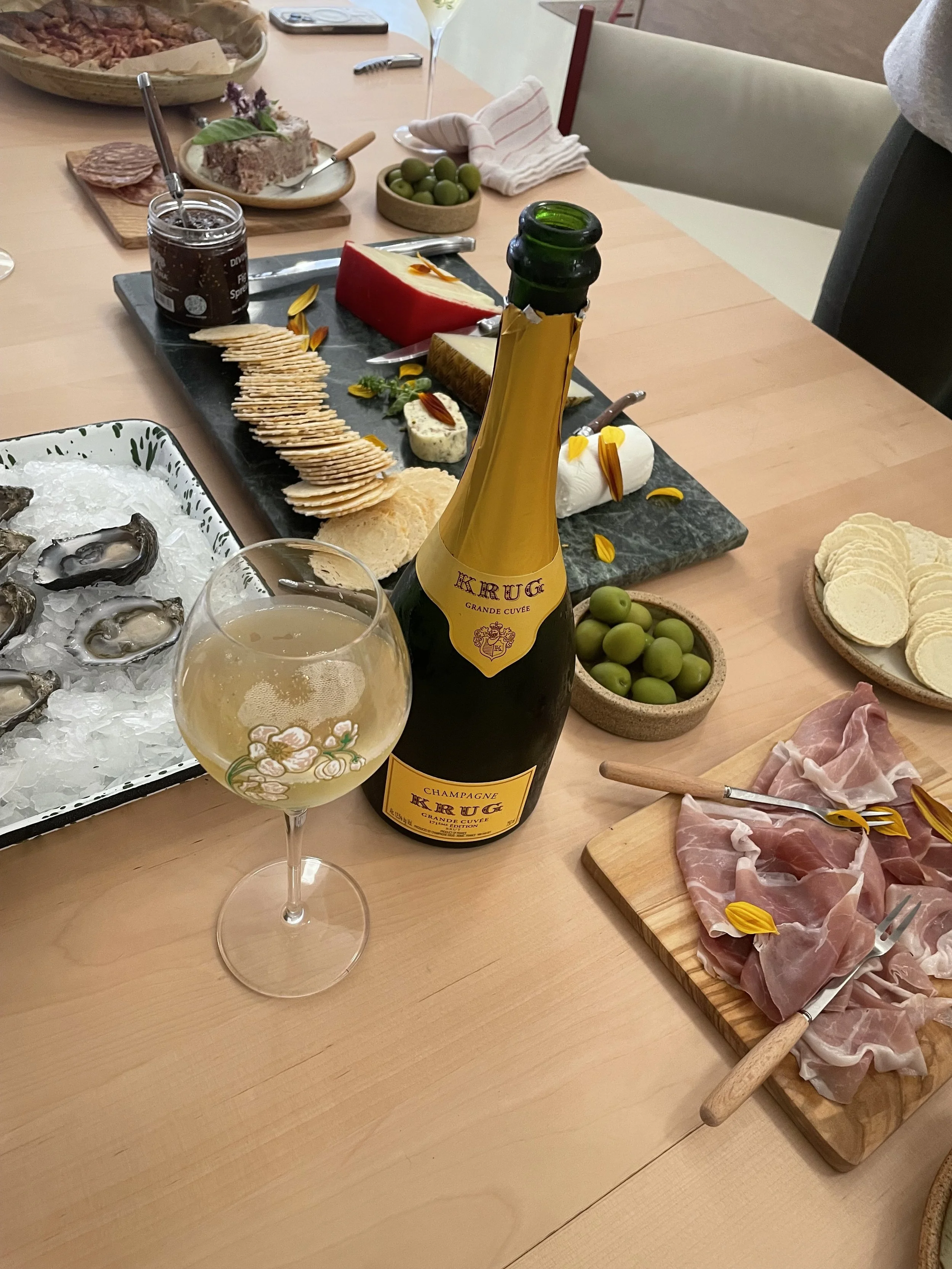 A table set with charcuterie, cheese, crackers, olives, oysters on ice, and a bottle of champagne, along with drinking glasses and various utensils and serving dishes.