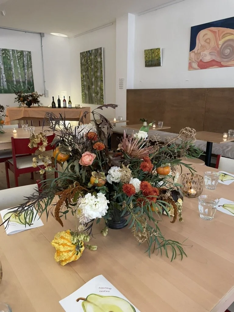 A floral centerpiece with autumn-colored flowers, small pumpkins, and greenery on a dining table set for a meal in a decorated restaurant.