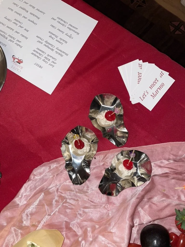 Three small desserts with cherries on top placed in reflective metallic bowls on a pink fabric, with a red tablecloth and cards reading 'meet at,' 'Let's meet at,' and 'Mumbo' nearby.