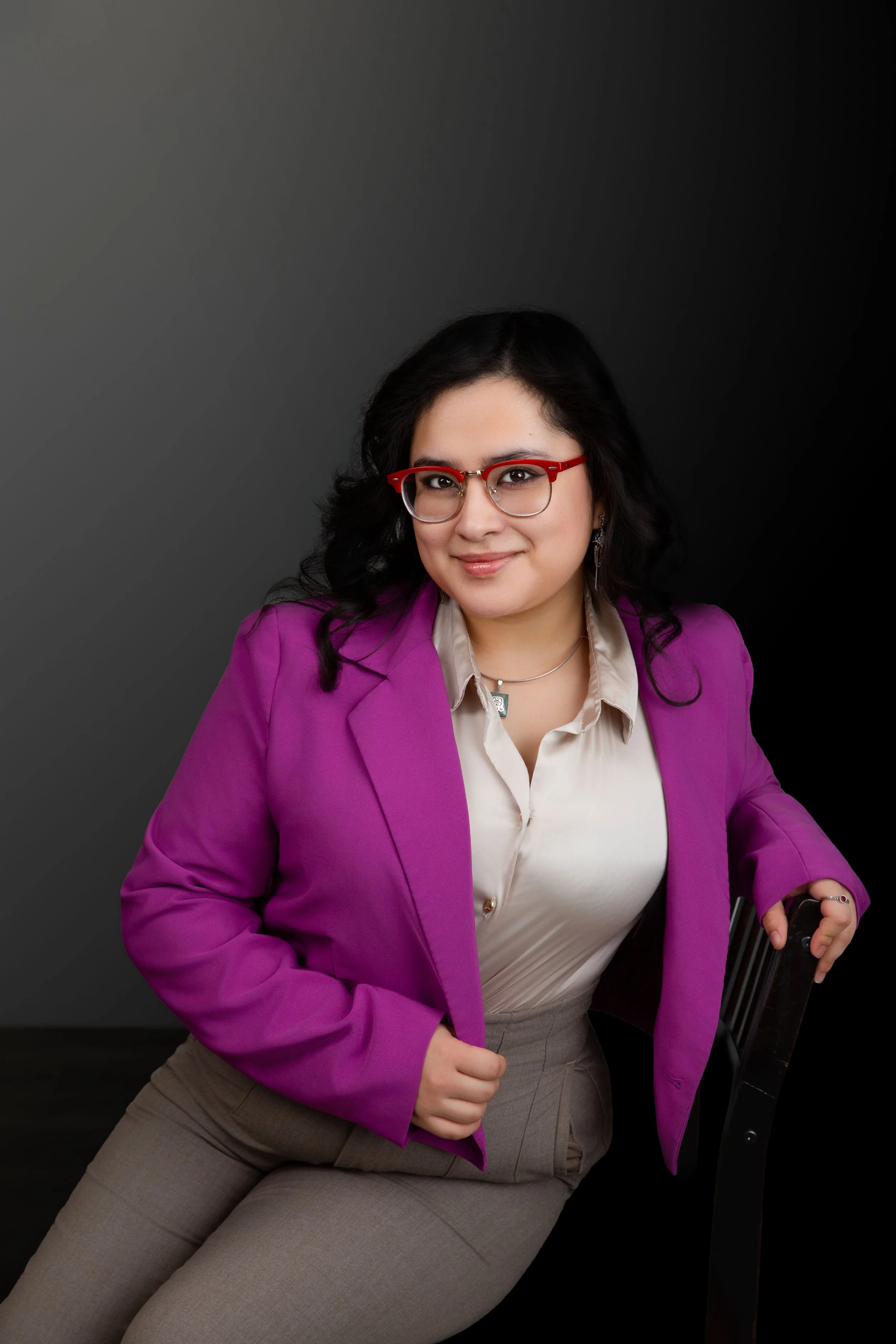 Andrea Cardenas, a woman with black hair, wearing glasses with red frames, a purple blazer, a beige shirt, and beige pants, sitting on a chair against a dark background.