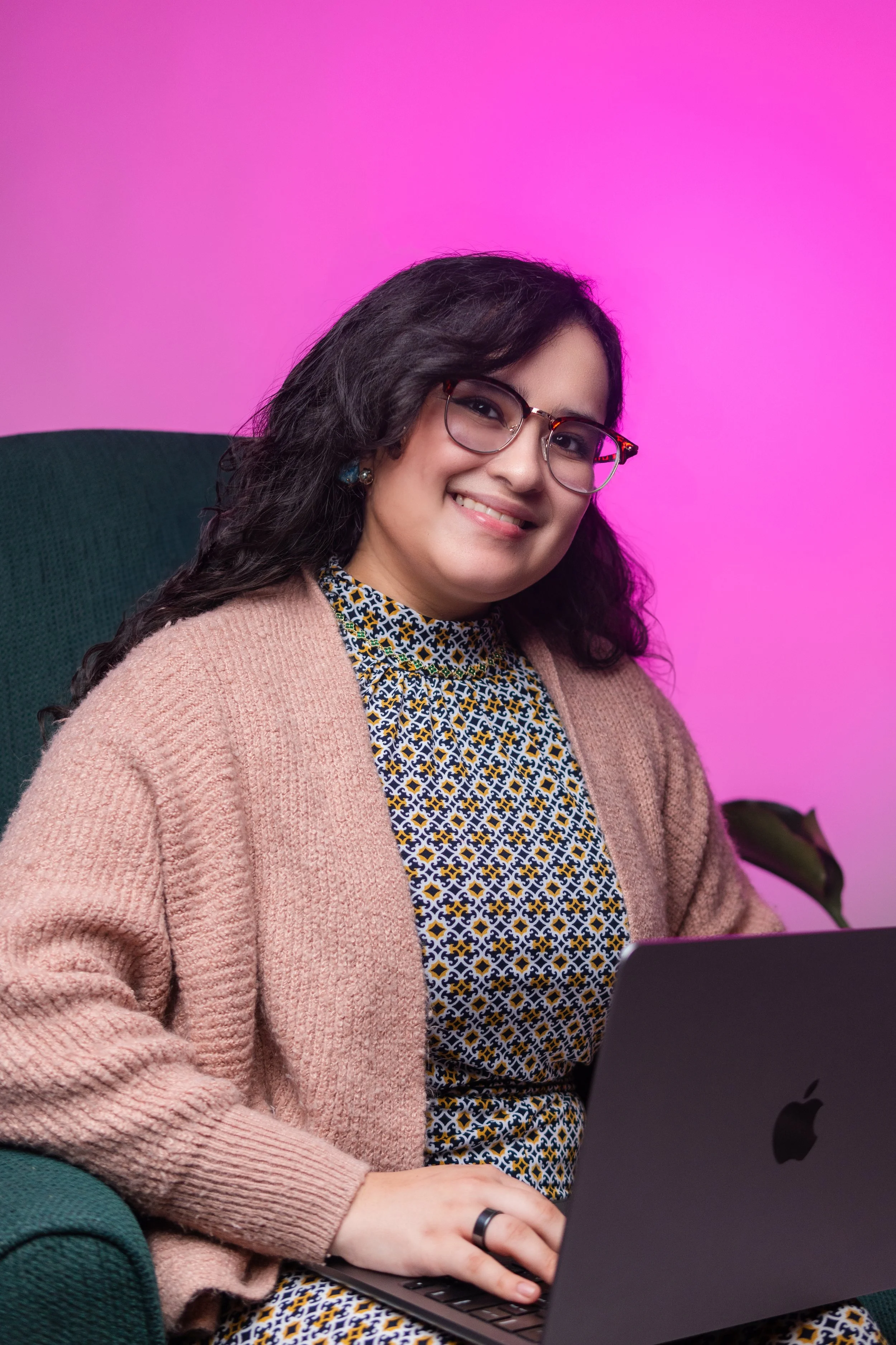 A woman with glasses sitting in a green chair using a laptop, with a pink gradient background.