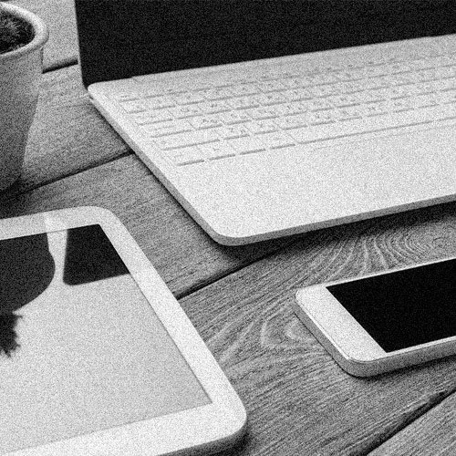 A wooden desk with a laptop, tablet, and smartphone on it, next to a plant in a pot.