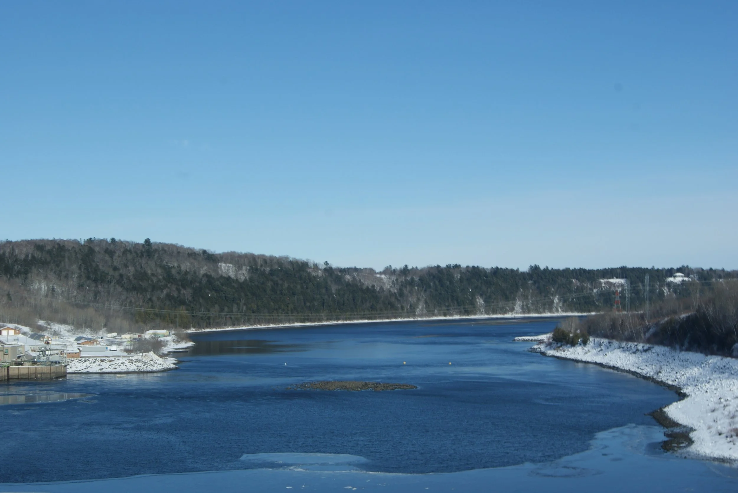 View of a river with some ice on the edges, snow on the banks, and wooded hills in the background under a clear blue sky.
