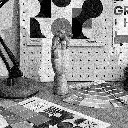 A wooden hand sculpture holding a small sphere, placed on a table with color swatches and design samples, in front of a geometric poster on a pegboard wall.