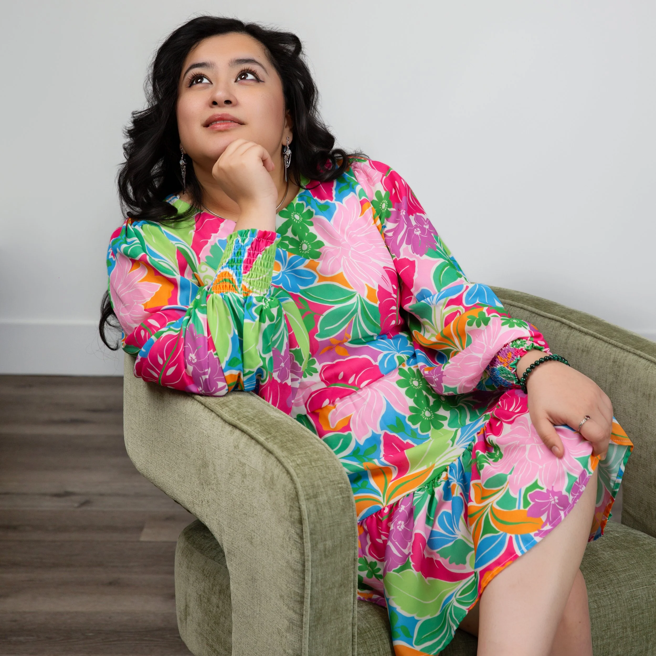 A woman in a colorful floral dress is sitting on a green chair with her right hand resting on her chin, looking upwards with a thoughtful expression.