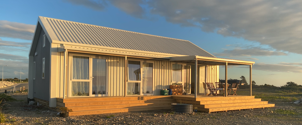 Modular modern home clad in light corrugated steel with gable roof, large sliding doors, and a covered timber deck catching the golden evening light in a coastal setting
