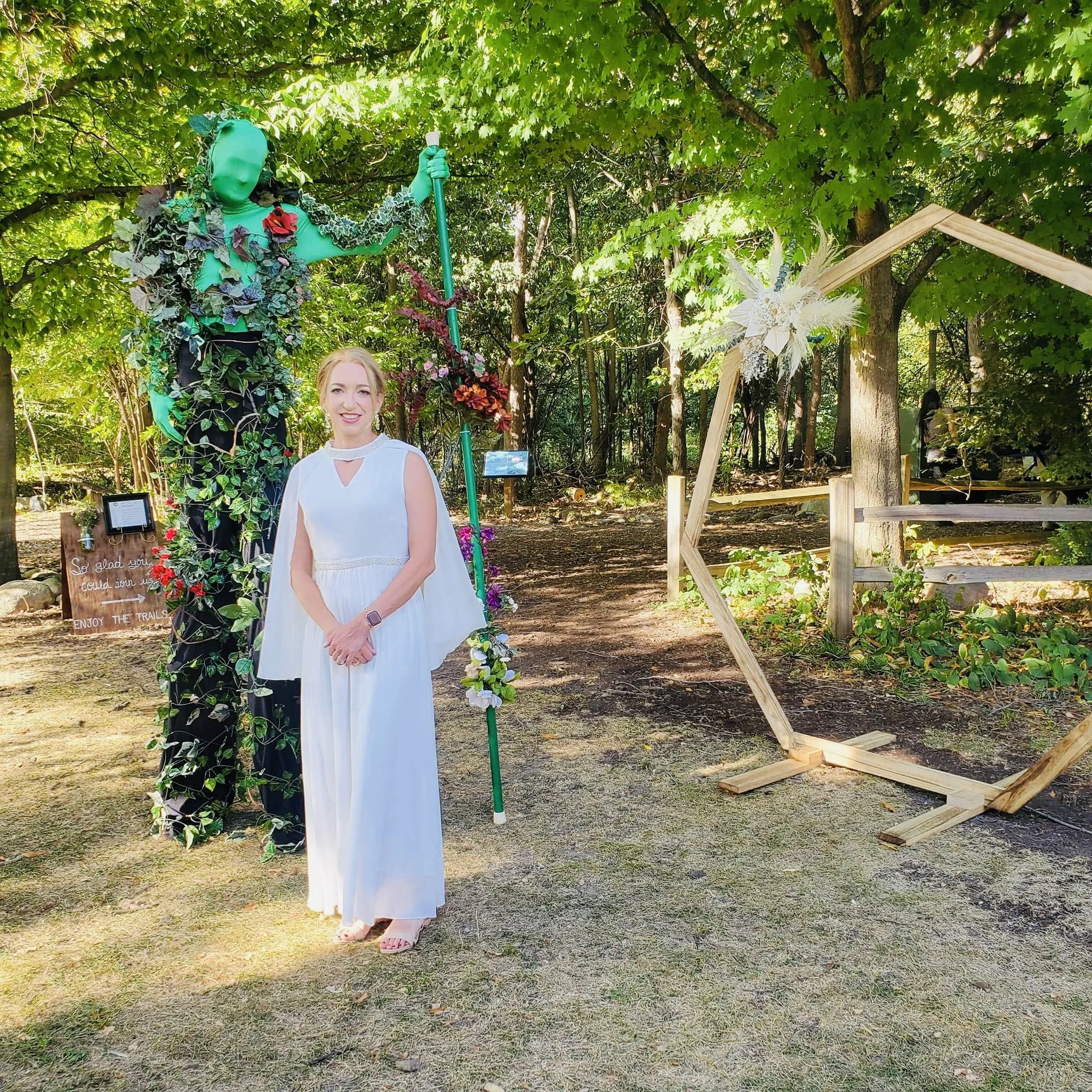 A woman in a white dress standing in a wooded outdoor area next to a large green humanoid statue covered in leaves and flowers, with a wooden geometric structure to the right.