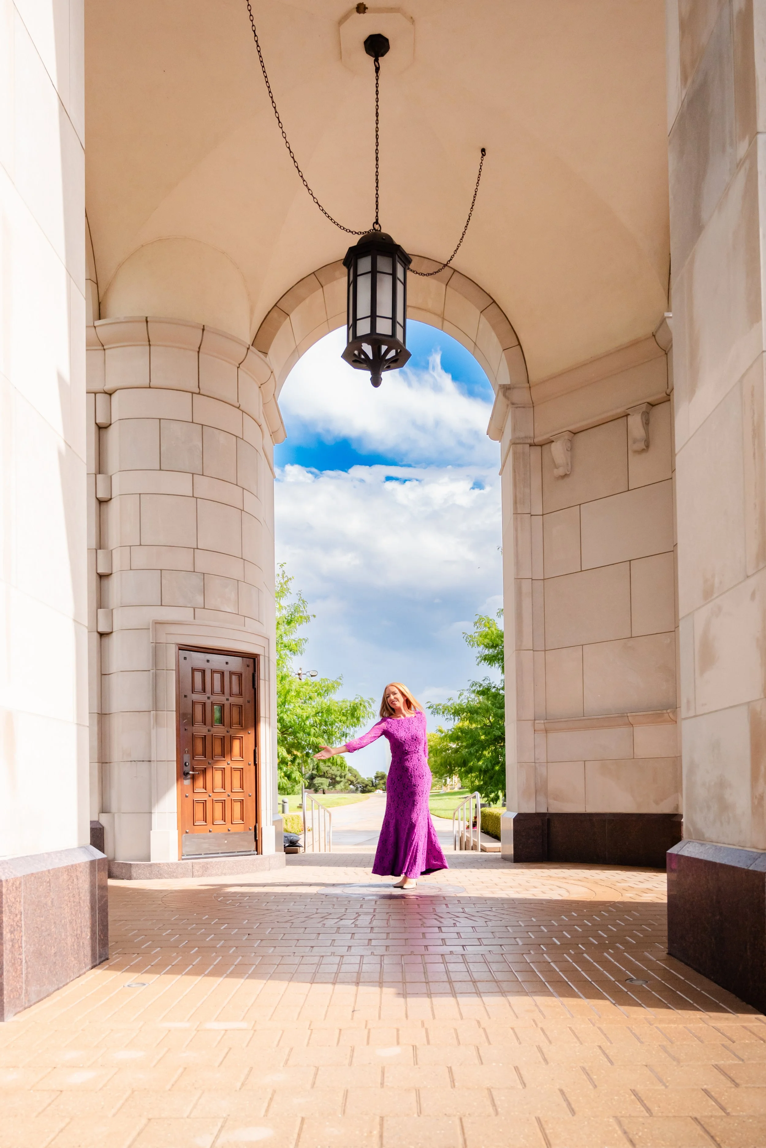 A woman in a purple dress dancing under an archway with a black hanging lantern, a cloudy blue sky, and green trees in the background.