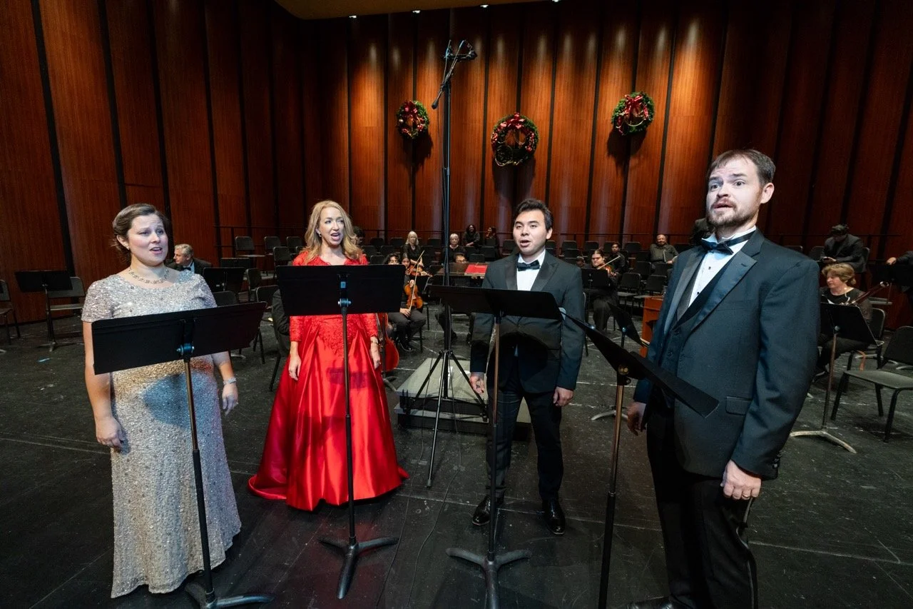 Four singers in formal attire performing on stage with an orchestra in the background, decorated with Christmas wreaths.