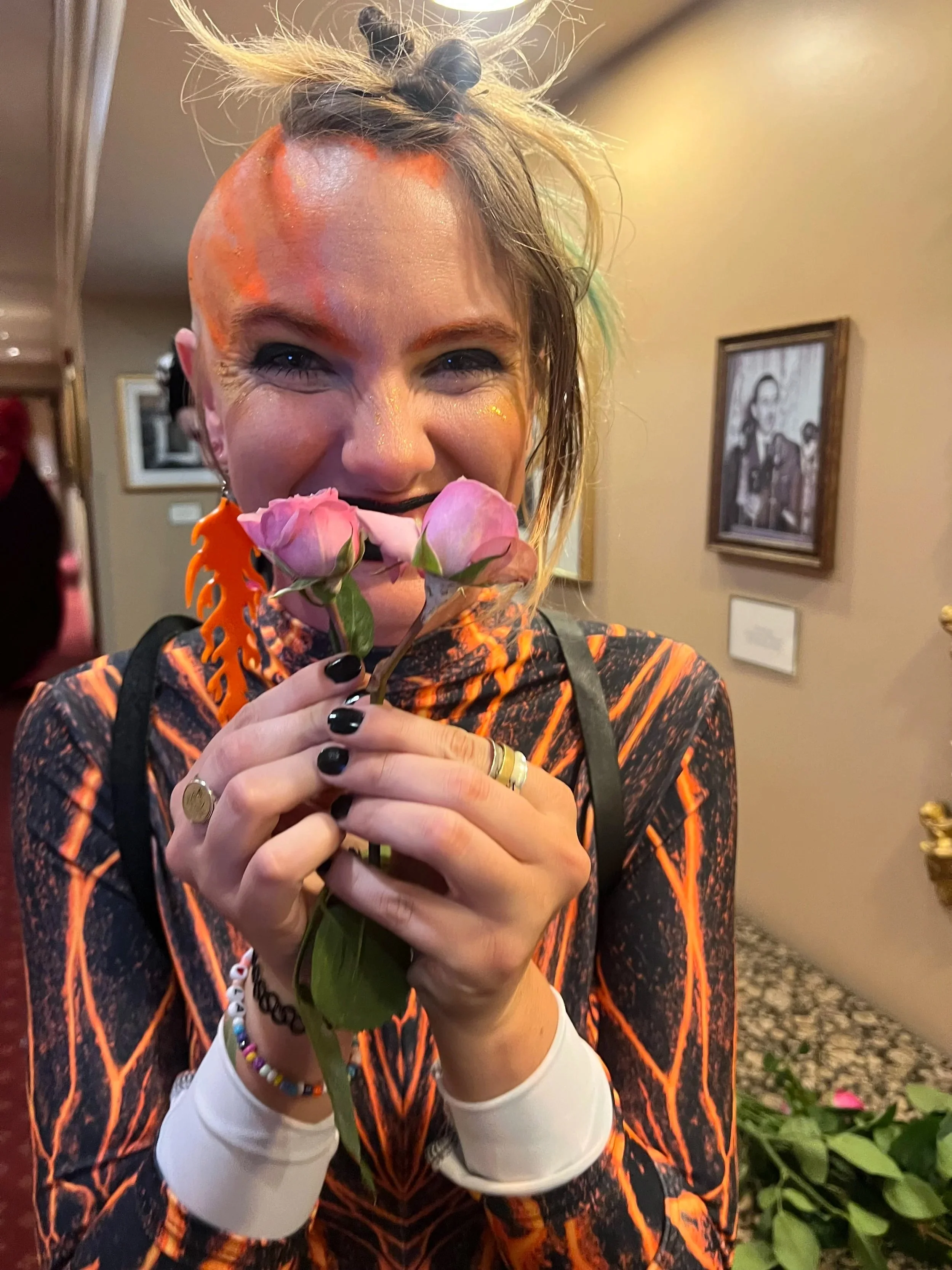 Asteria is dressed in her Fire Demon Costume for Labyrinth Masquerade, holding two tiny pink roses over their mouth. Their nails are black and their eyes are smiling.