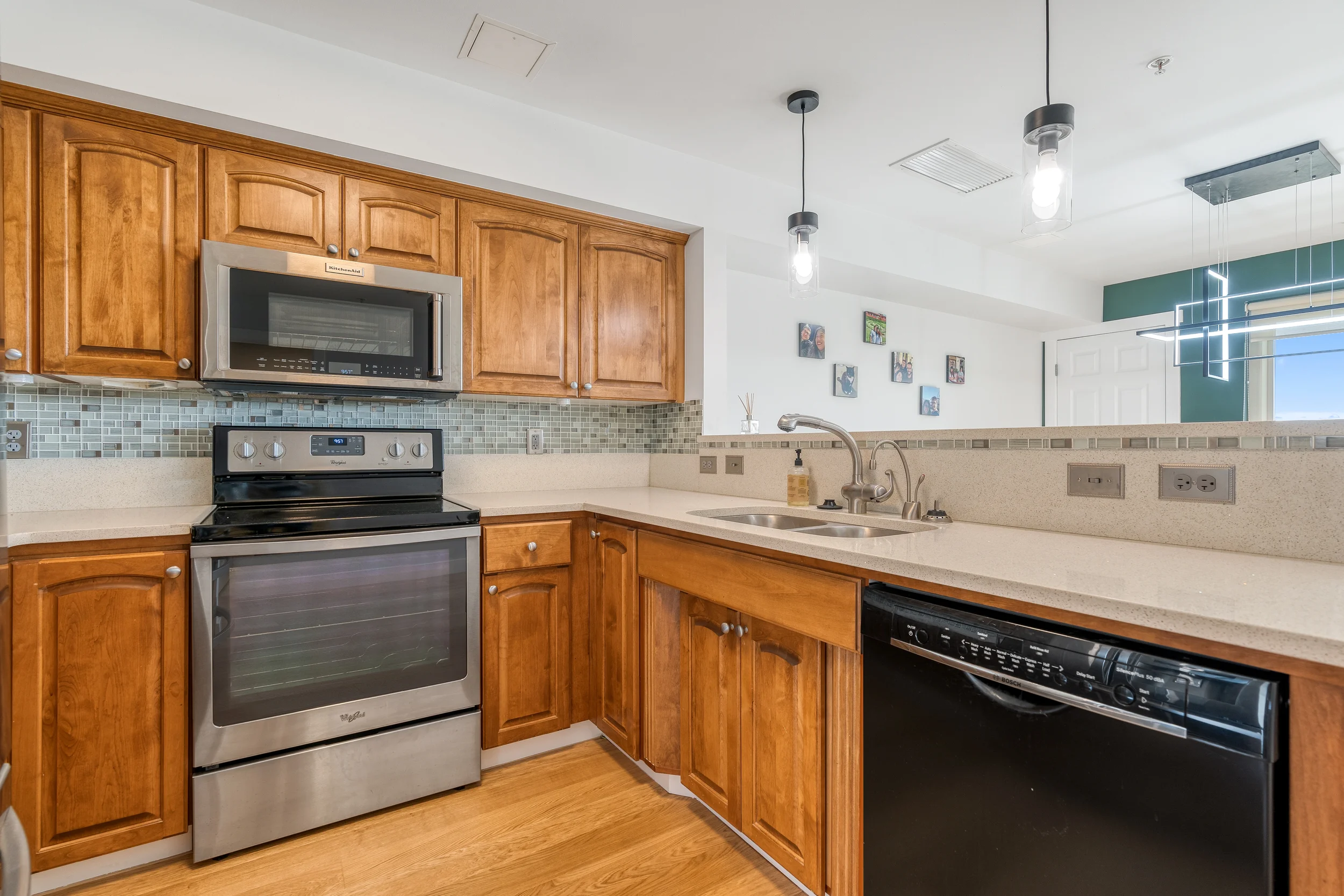 Kitchen with wooden cabinets, a stainless steel microwave and oven, a black dishwasher, a beige countertop, a stainless steel sink, a soap dispenser, hanging light fixtures, and a wall with family photos and electrical outlets.