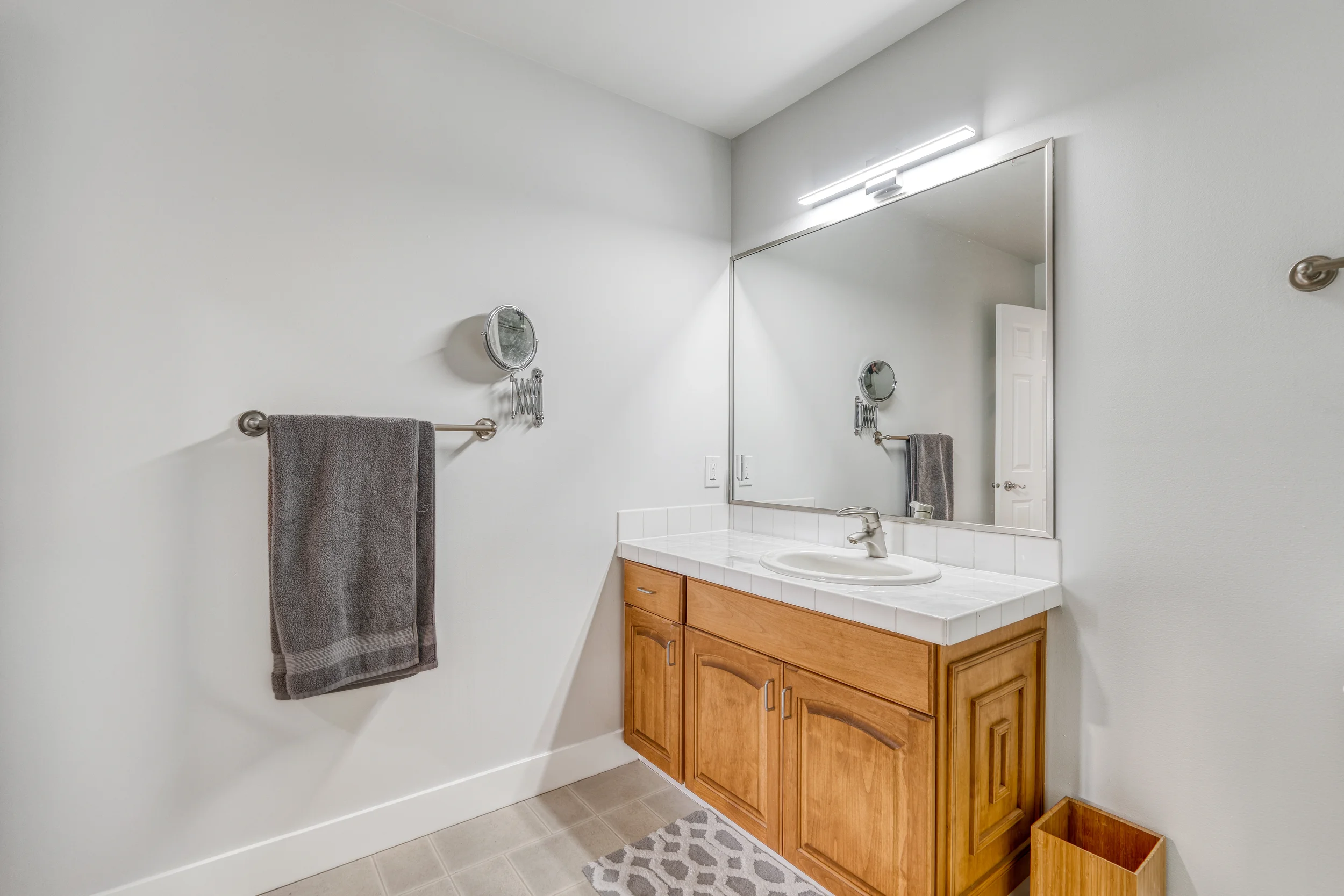 Bathroom with a wooden vanity, white tiled countertop, large mirror, gray towel on a bar, small round mirror, light fixture above the mirror, light-colored wall, electrical outlet, and a door on the right side.