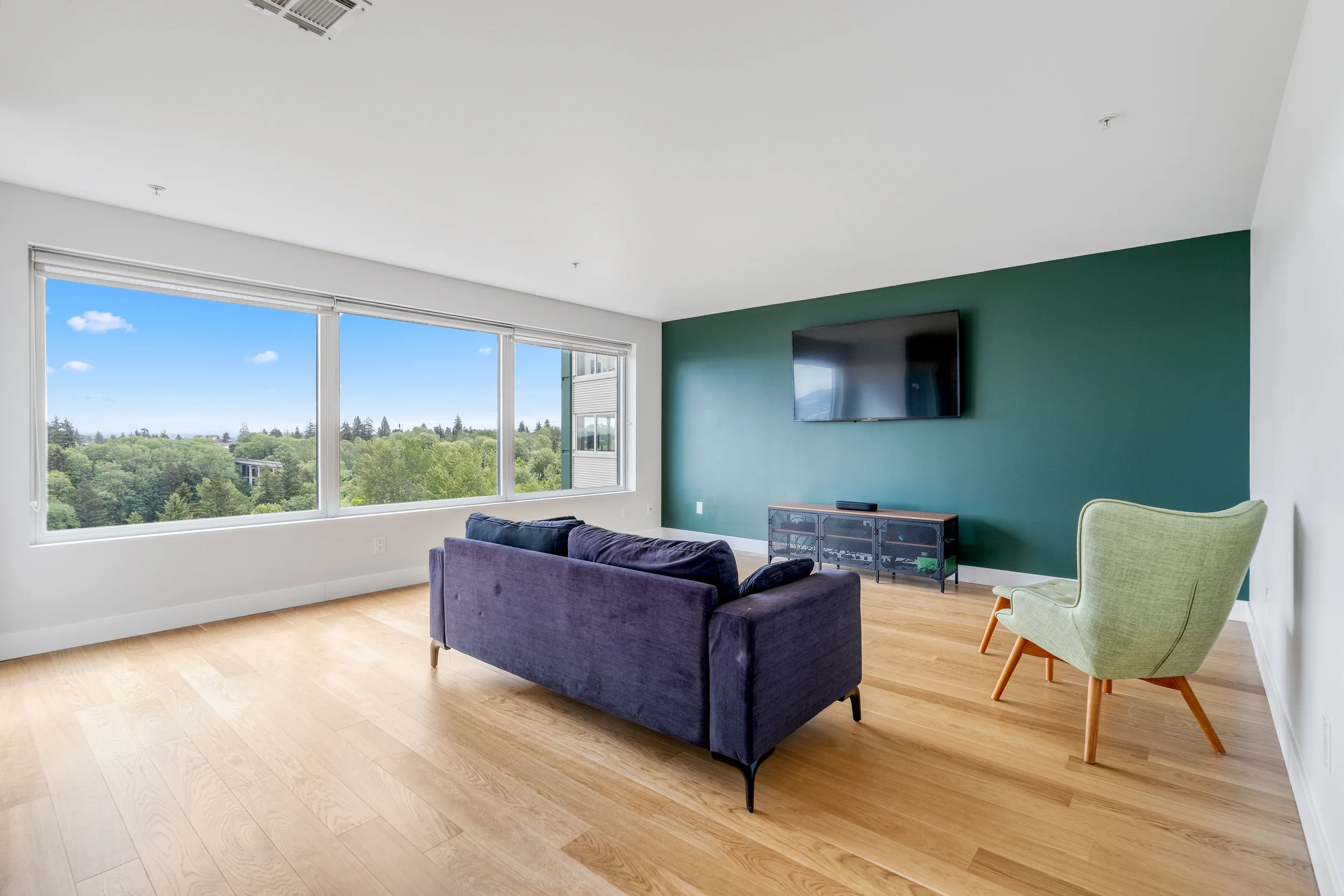 A modern living room with large windows showing a green, tree-filled landscape and a clear blue sky, a purple sofa, a light green armchair, a dark wood TV stand, a flat-screen TV on a dark green accent wall, and hardwood flooring.