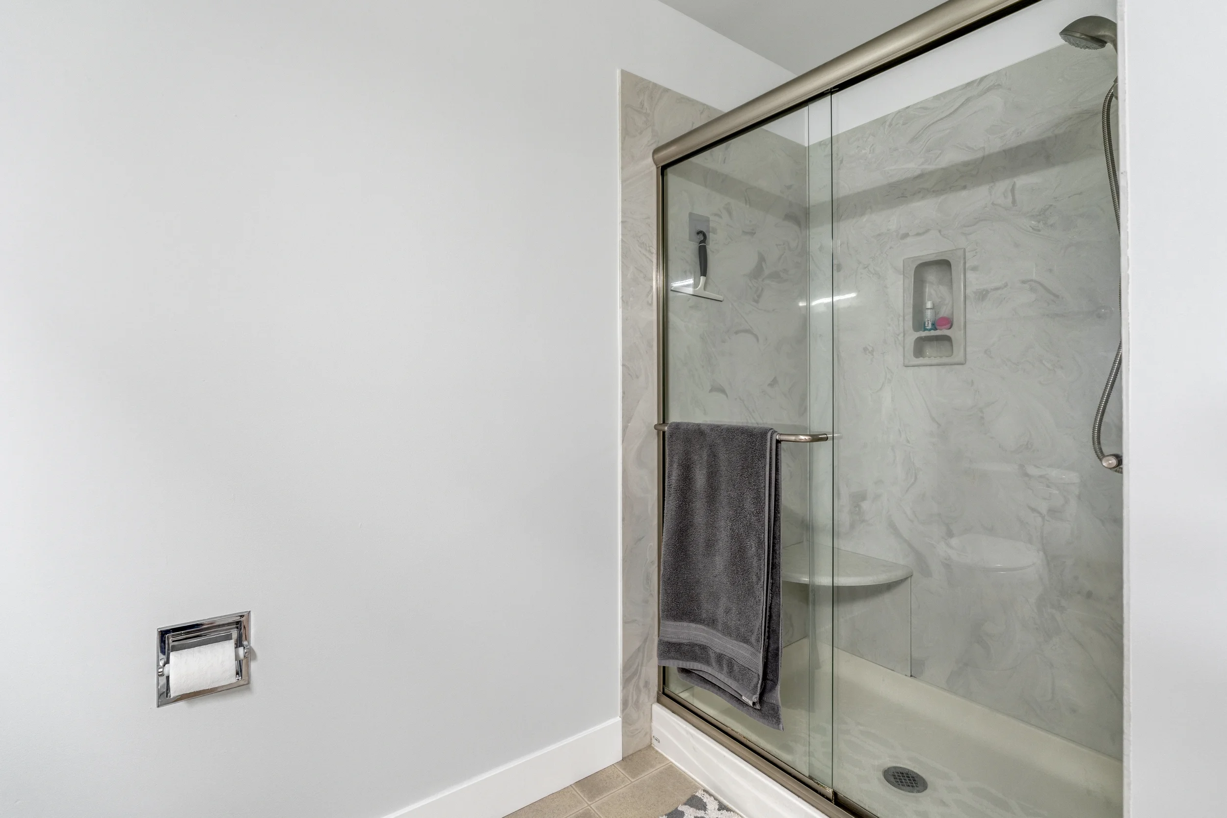 A modern bathroom shower with glass doors, a gray towel hanging on the handle, a built-in shelf with toiletries, and a marble wall.