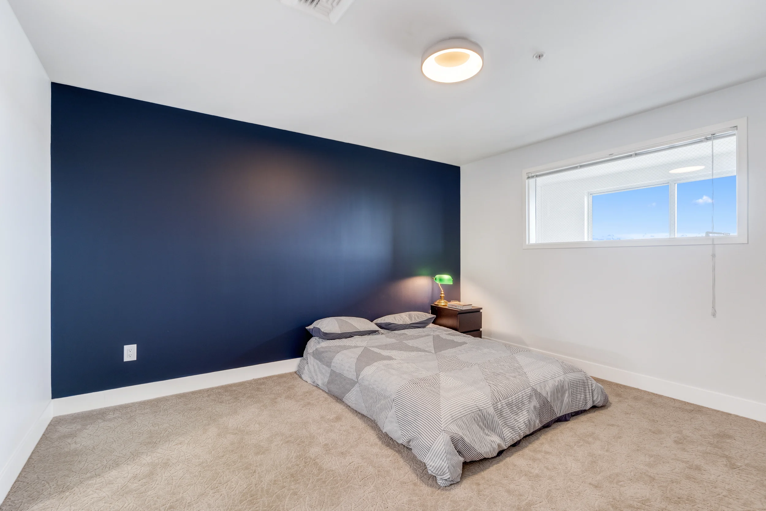 A minimalist bedroom with a dark blue accent wall, a bed with gray and white bedding on the floor, a small wooden nightstand with a green desk lamp, and a window with a view of the sky.