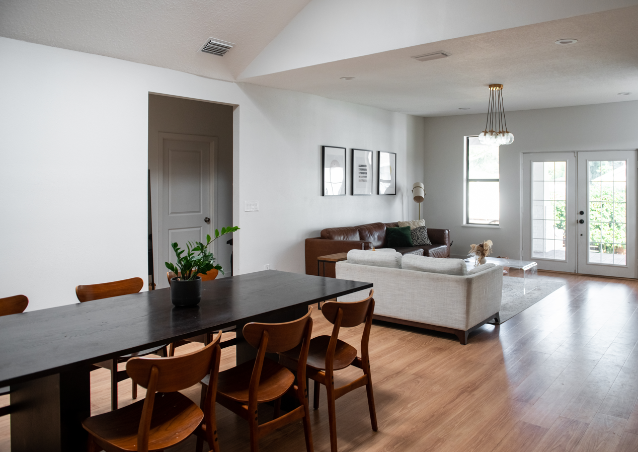 Living room with wooden flooring, a white sofa, a brown leather sofa, a glass coffee table, a window, and background artwork.