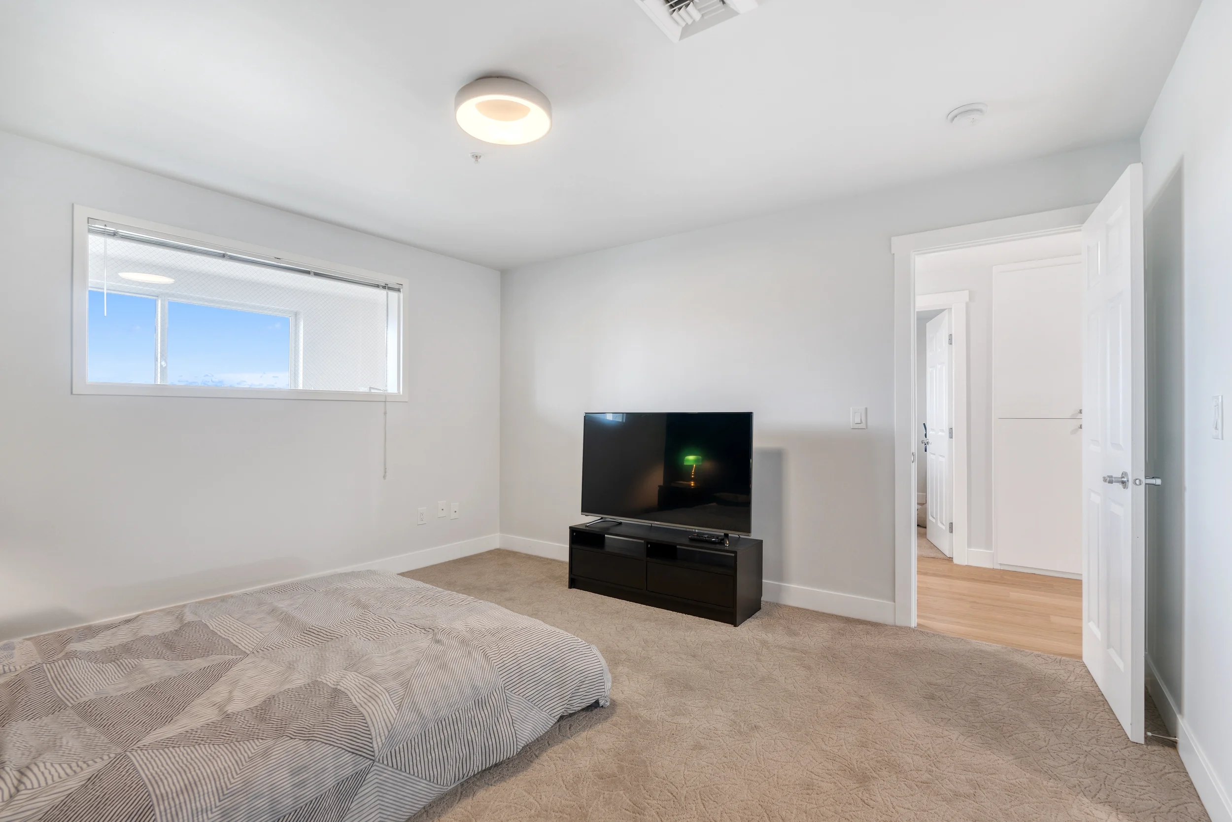 A living room with a bed, a small black TV stand with a TV, a large window, and open doorways to other rooms, beige carpet, white walls, and minimal furniture.