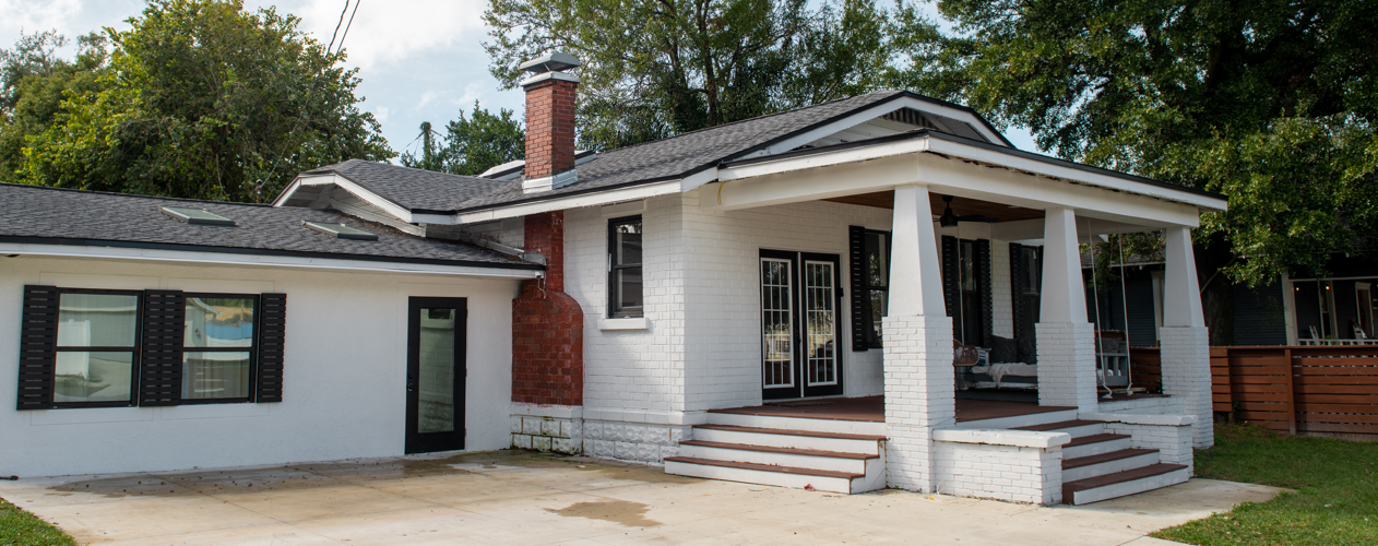 White brick house with black shutters, a porch with steps, a swing, and a chimney, surrounded by trees and a wooden fence.