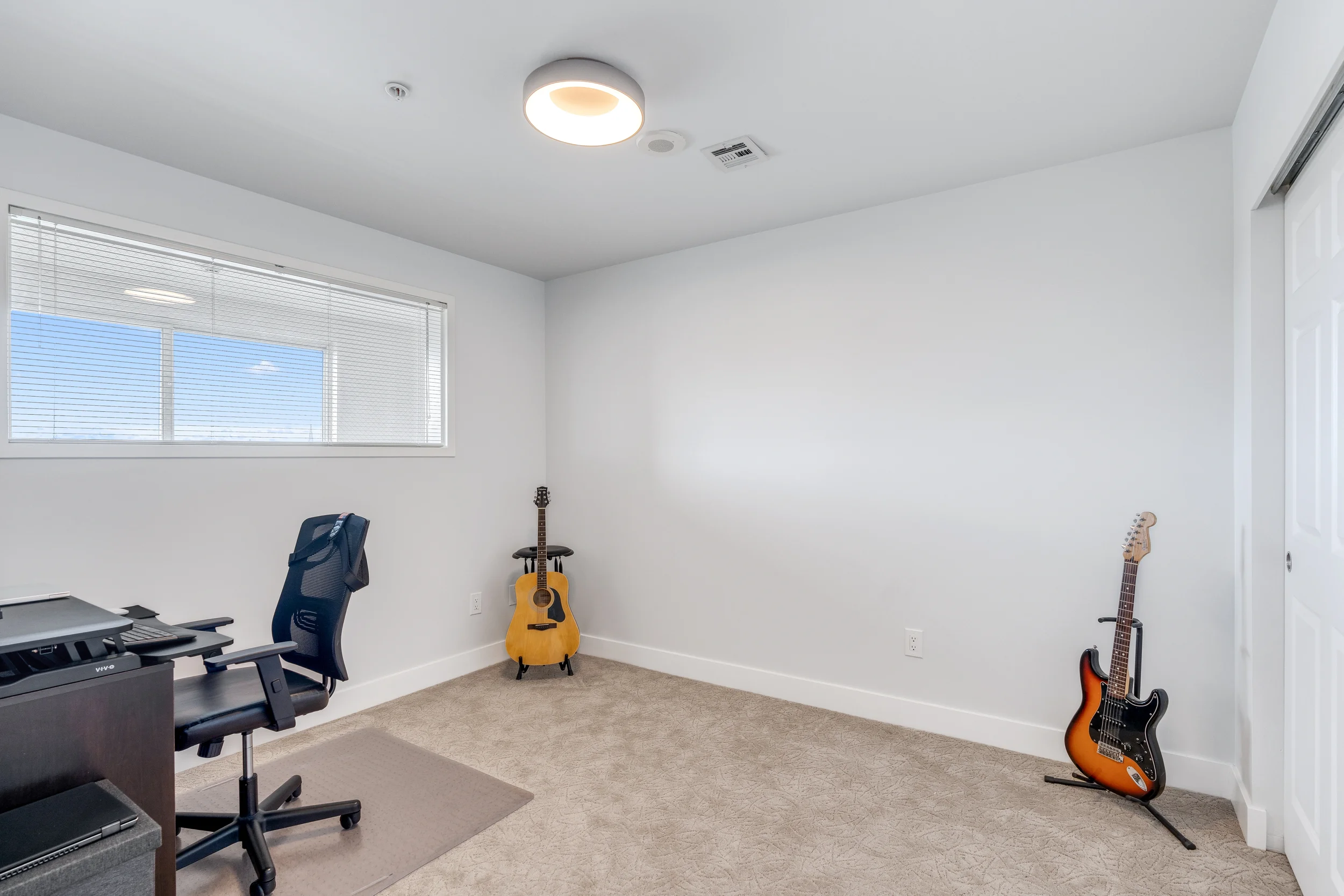 A room with white walls, beige carpet, a window with blinds, a black office chair, a desk with a printer, two guitars (an acoustic and an electric), and a ceiling light fixture.