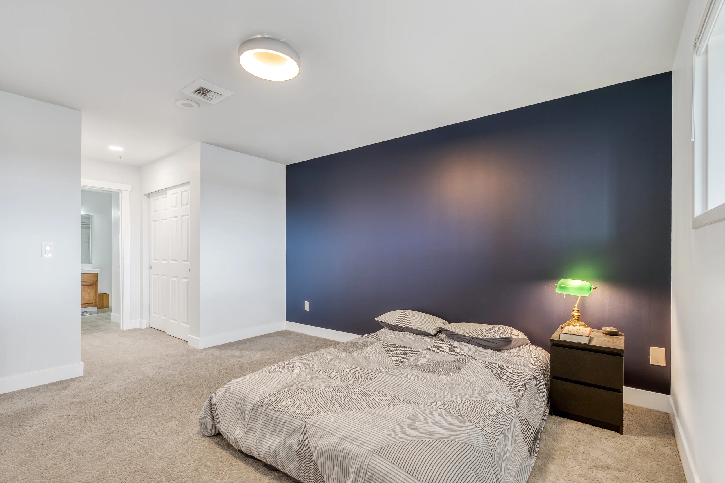 A bedroom with a bed in the foreground, a nightstand with a green lamp, and a blue accent wall. There is a small window on the right and a white ceiling with a ceiling light. The room has beige carpet.