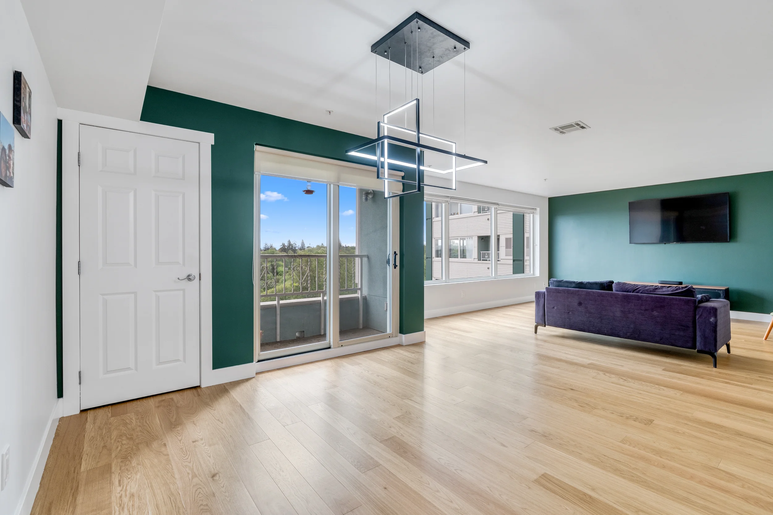 Modern living room with hardwood floors, green accent wall, large windows, a purple sofa, and a unique black chandelier with geometric shapes.