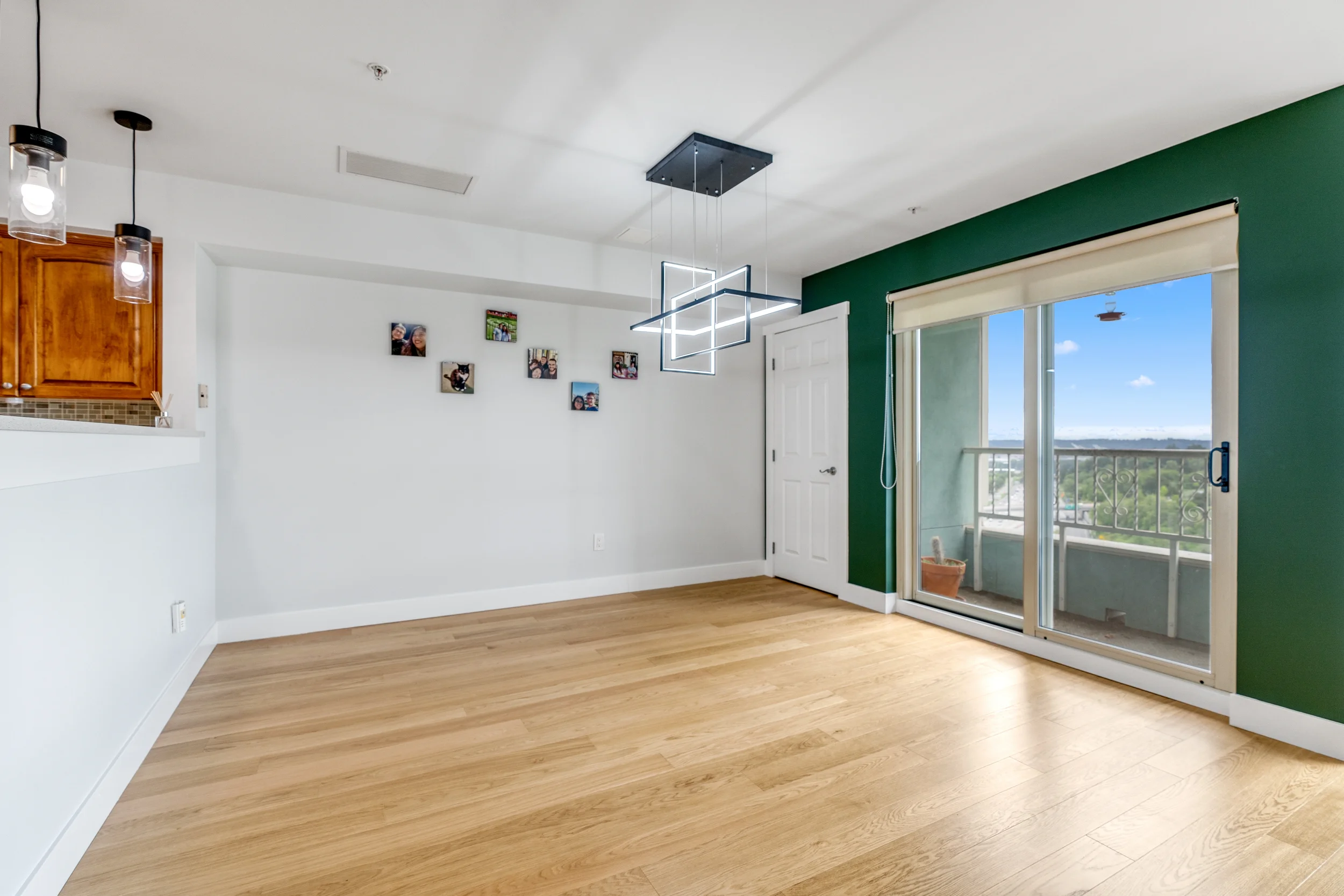 A bright living room with hardwood floors, a large sliding glass door leading to a balcony, a green accent wall, white walls, a modern rectangular LED ceiling light, and a wall decorated with family photos.