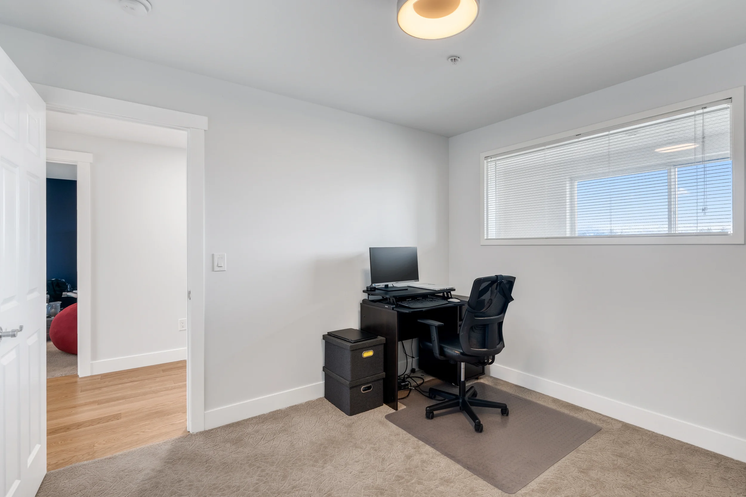 A small home office with a black desk, a monitor, and an office chair, next to a large window with blinds. The room has a beige carpet and white walls.