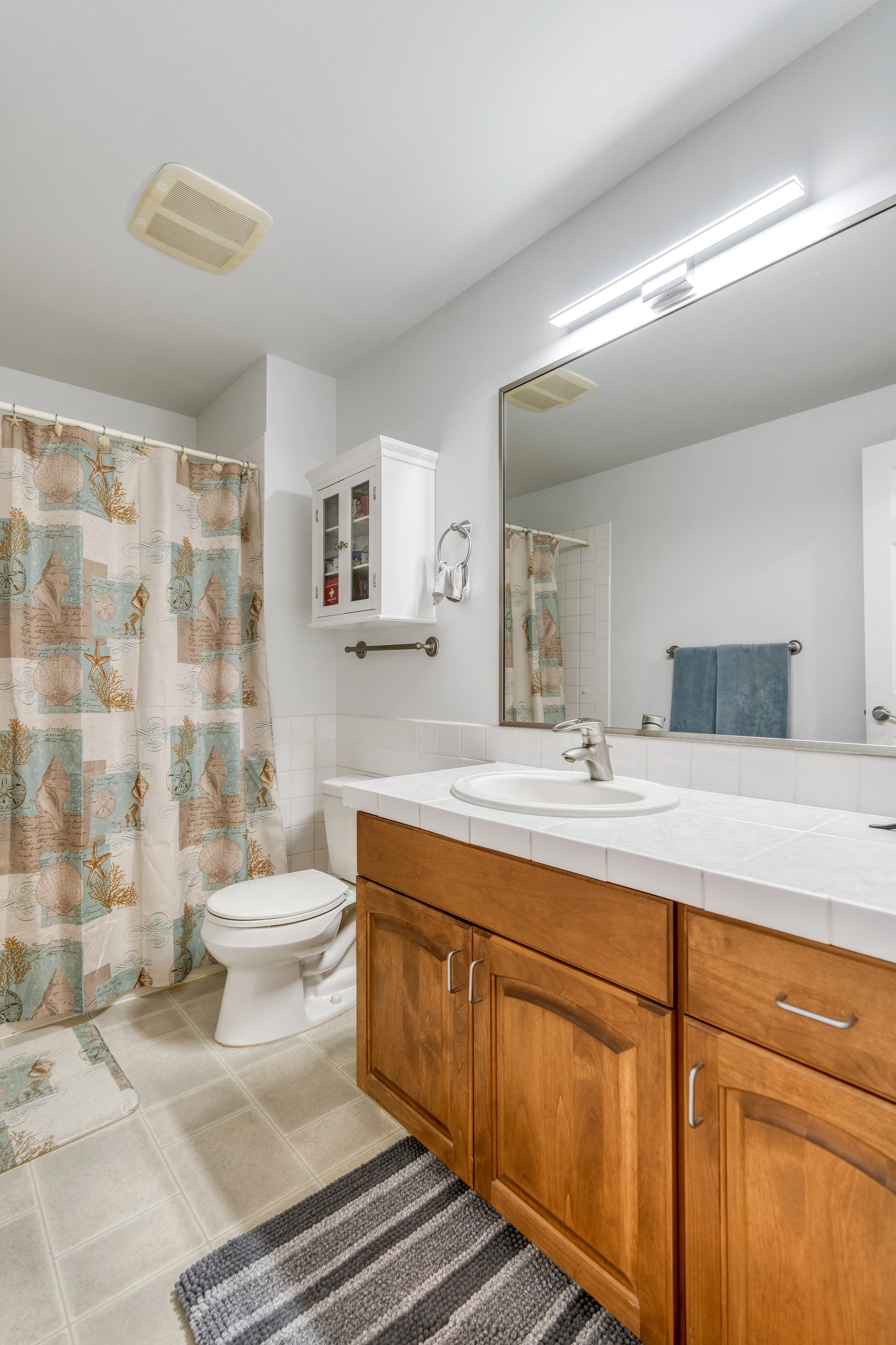 A bathroom with a shower curtain with seashell and starfish design, a toilet, a white wall cabinet, a mirror with an overhead light fixture, a sink with a wooden cabinet, a towel rack with a blue towel, a black and gray striped rug on beige tile floo