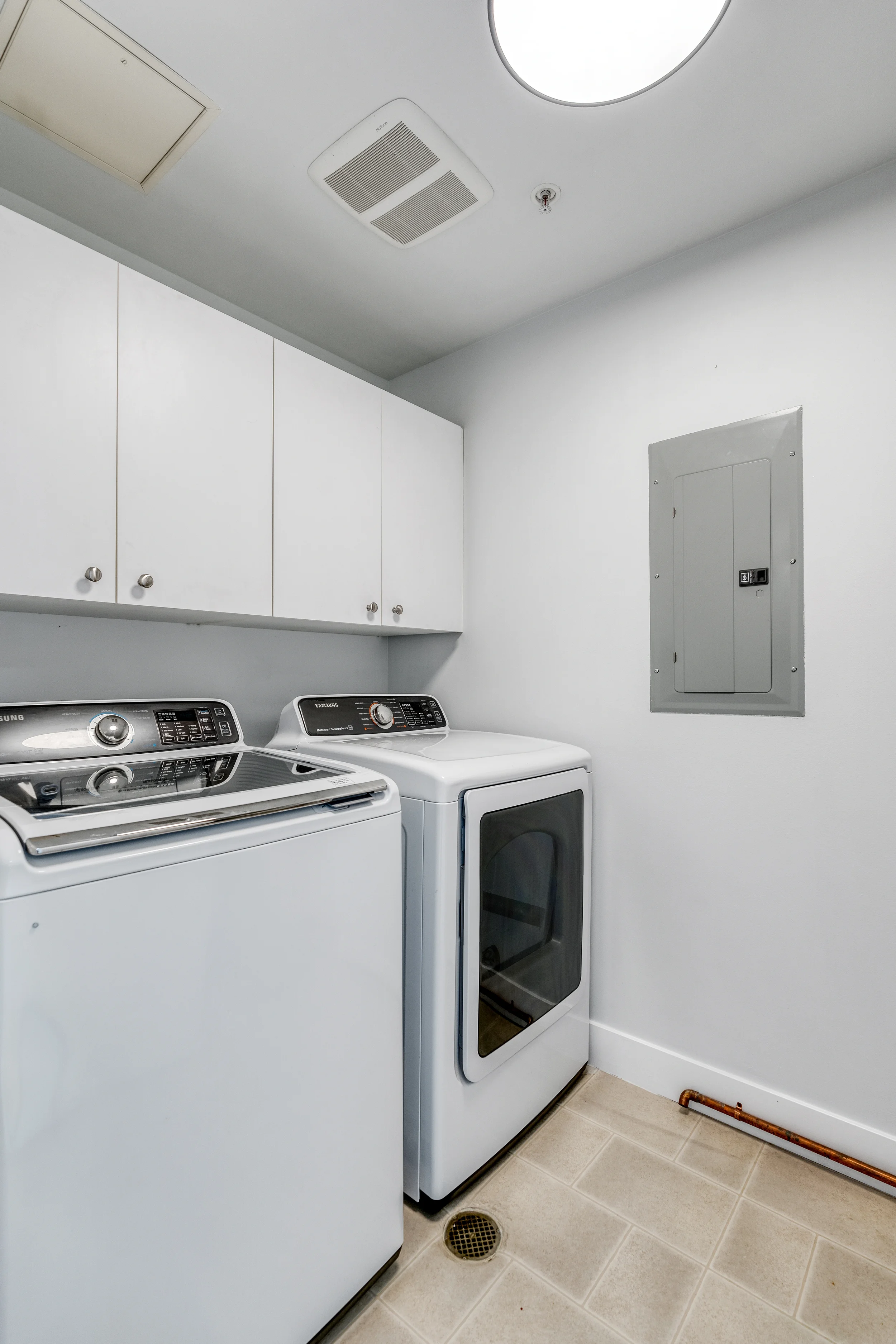 A laundry room with white cabinets and a white washing machine and dryer. The walls are white, and there is a gray electrical panel on the wall. The floor has beige tiles, and there is a round floor drain.