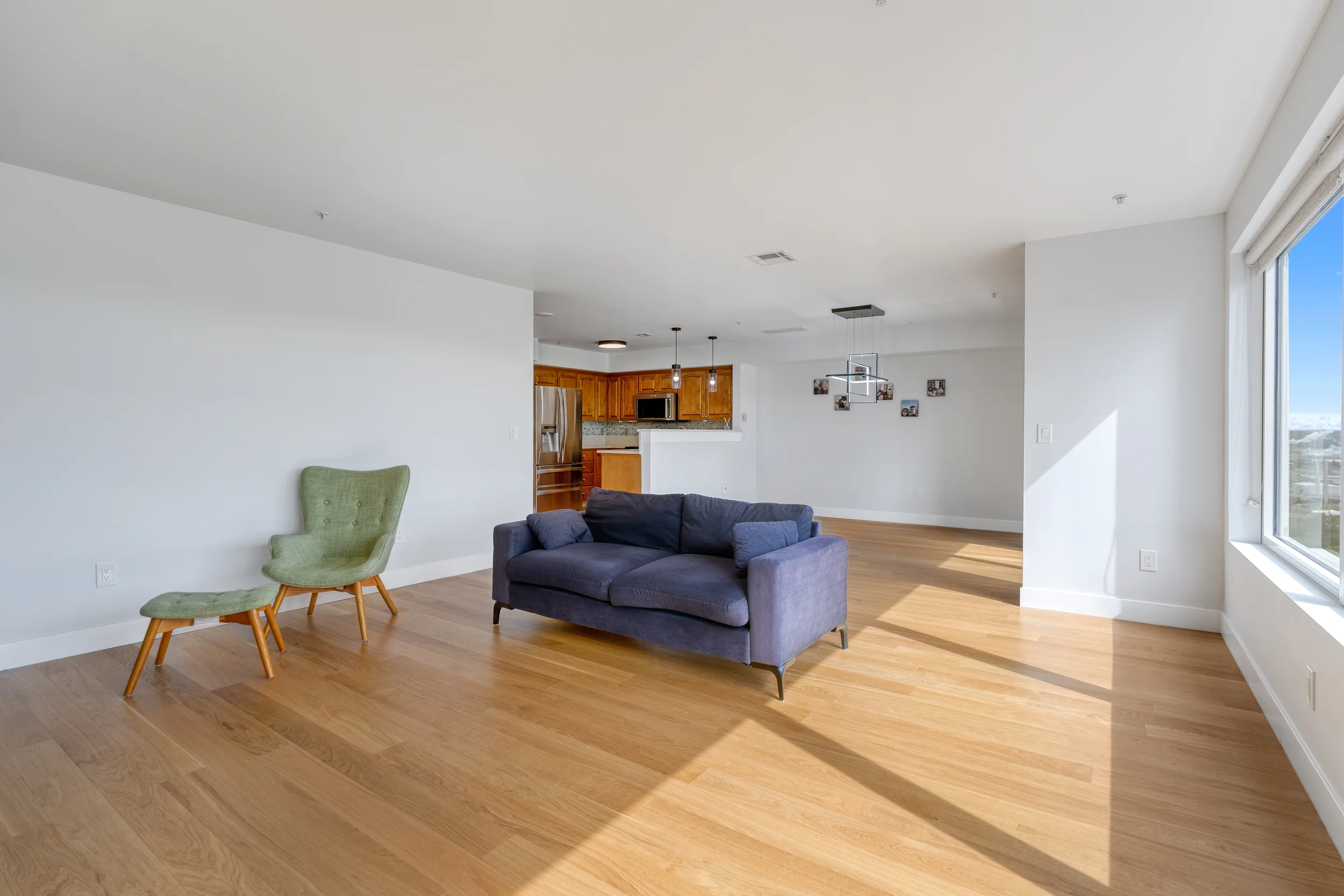 Living room with hardwood floors, blue sofa, green armchair, large window, and open kitchen with wooden cabinets and stainless steel appliances.