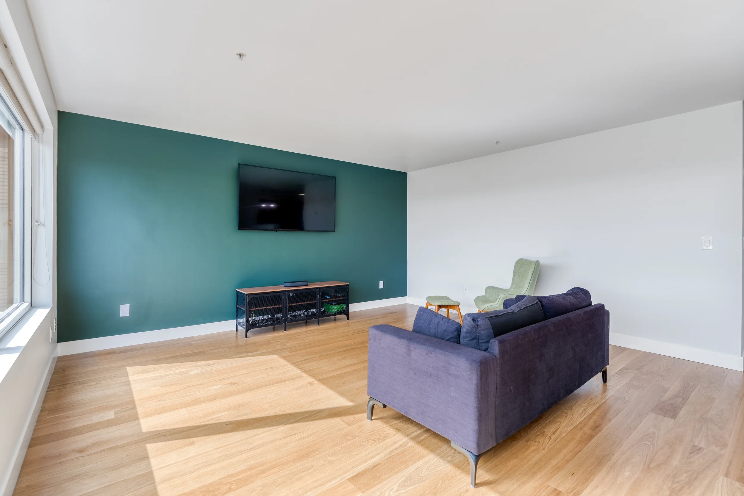 Living room with a dark purple couch, light green armchair, and a light green footstool. A large teal accent wall holds a wall-mounted flat-screen TV and a black and wooden media console. The room has hardwood floors and a large window on the left.