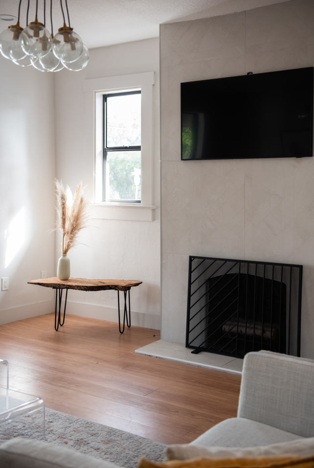 A living room corner with a white wall, a window, a small wooden table with black hairpin legs and a beige vase with pampas grass, a flat-screen TV mounted above a black fireplace grate, wooden flooring, and a portion of a light-colored armchair.