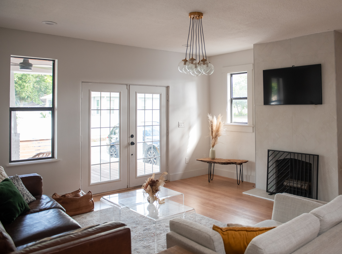 Living room with a leather sofa, a light-colored armchair, glass coffee table, and a velvet pillow. French doors leading outside, a window, a mounted flat-screen TV, a fireplace with a black grate, and a side table with a vase of pampas grass. Modern