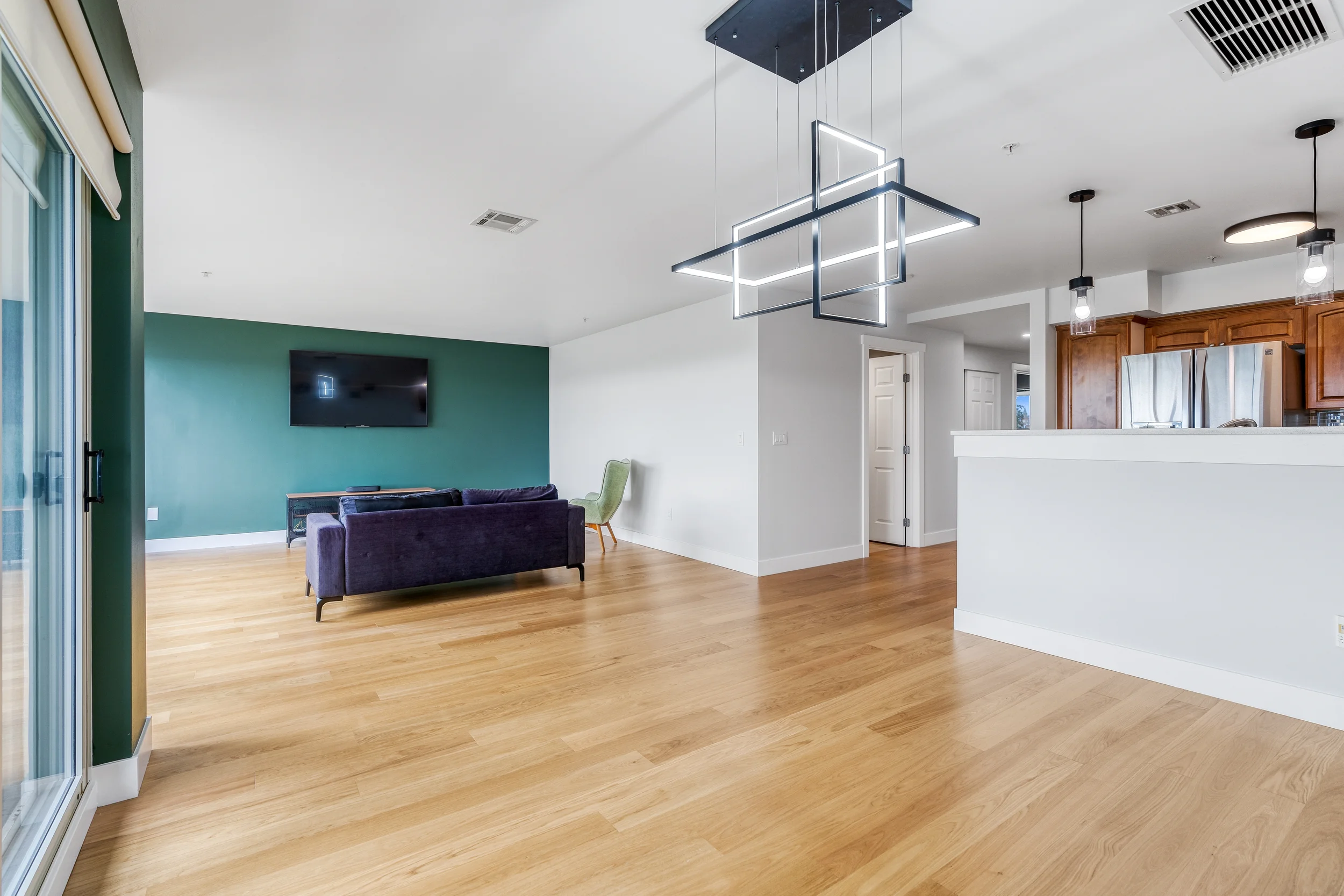 Living room with a purple sofa, a green armchair, a wall-mounted TV, a modern geometric light fixture, and wooden flooring.