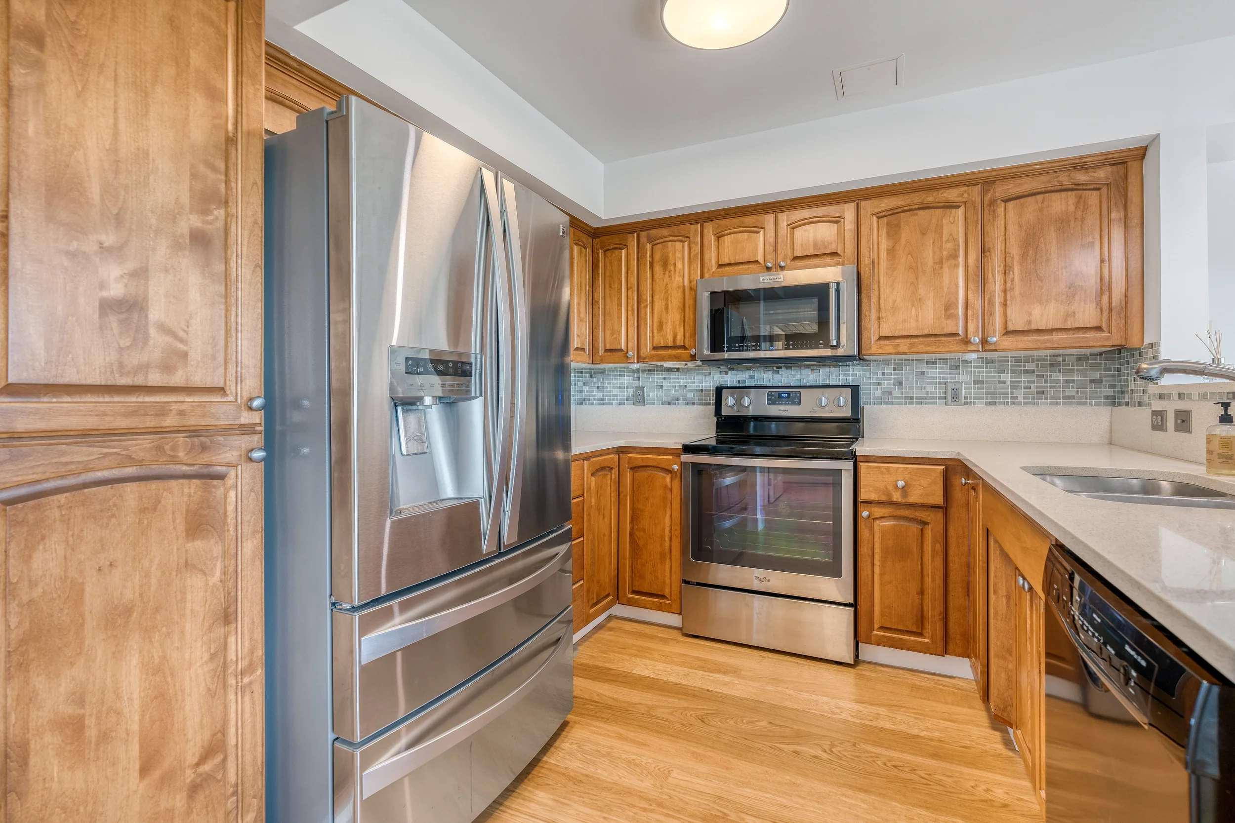 A kitchen featuring wooden cabinets, stainless steel appliances including a refrigerator, microwave, and oven, with a mosaic tile backsplash and beige countertops.