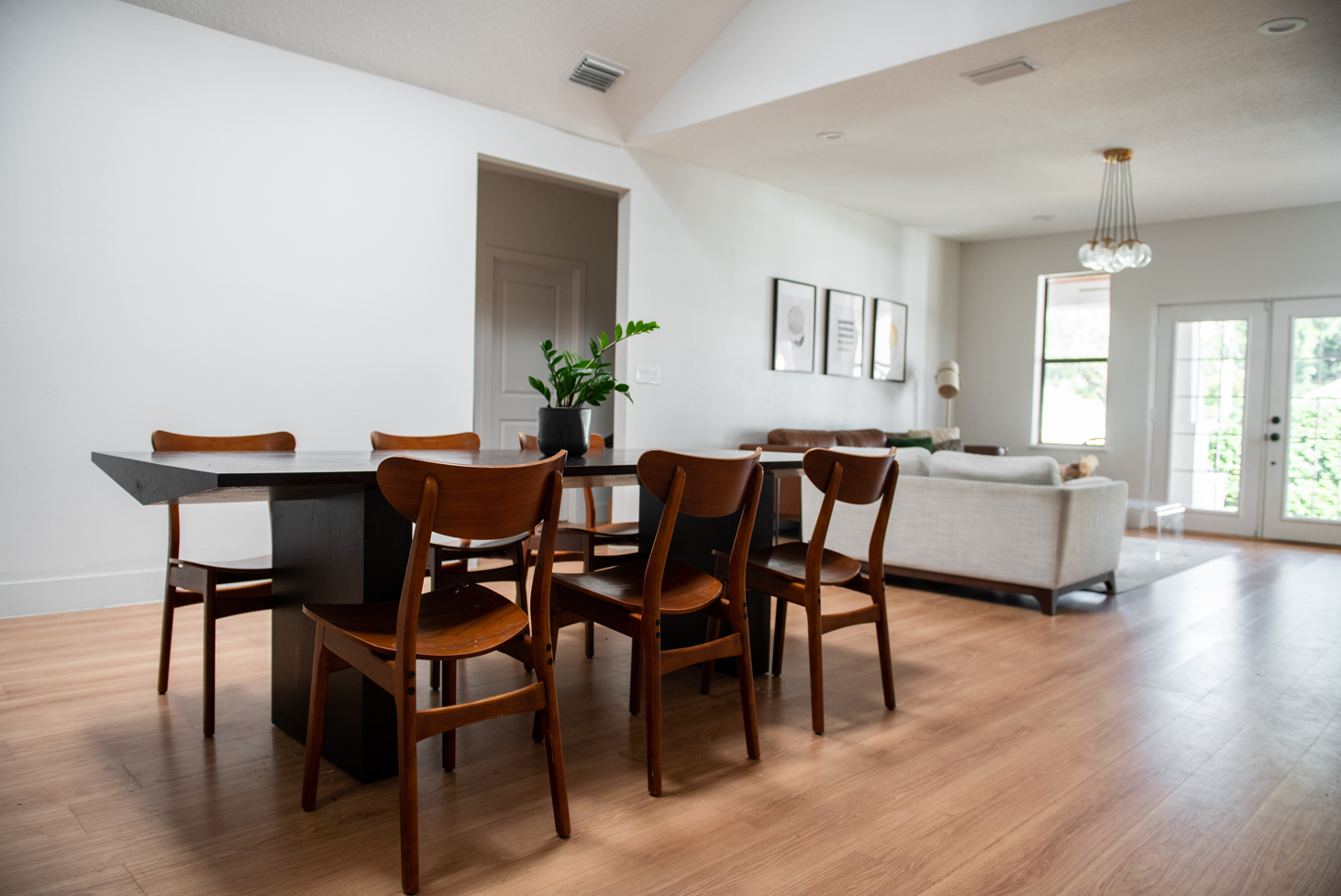 Interior of a modern living and dining room with a wooden dining table, six matching chairs, a white sofa, a beige armchair, framed wall art, a plant in a black pot, and large windows with glass doors.
