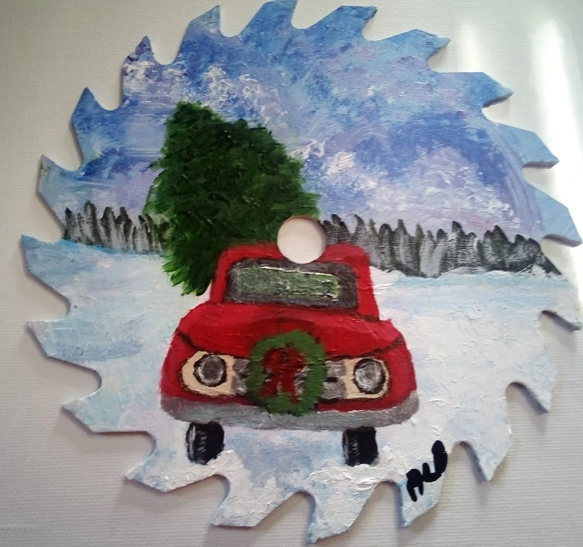 A painted winter scene on a big white snowflake-shaped cutout. It features a red truck with a wreath on the front, a green tree in the truck bed, a snowy background, and a blue sky.