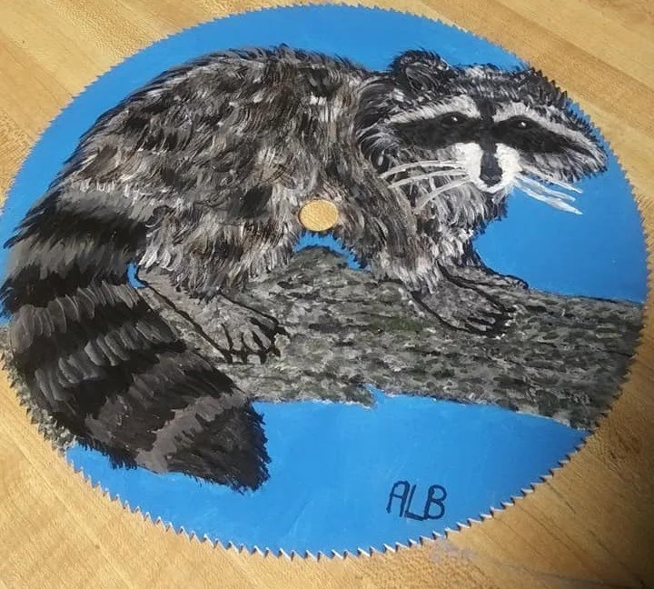 Painted circular wall art of a raccoon perched on a tree branch against a blue background, with the initials 'ALB' written at the bottom.